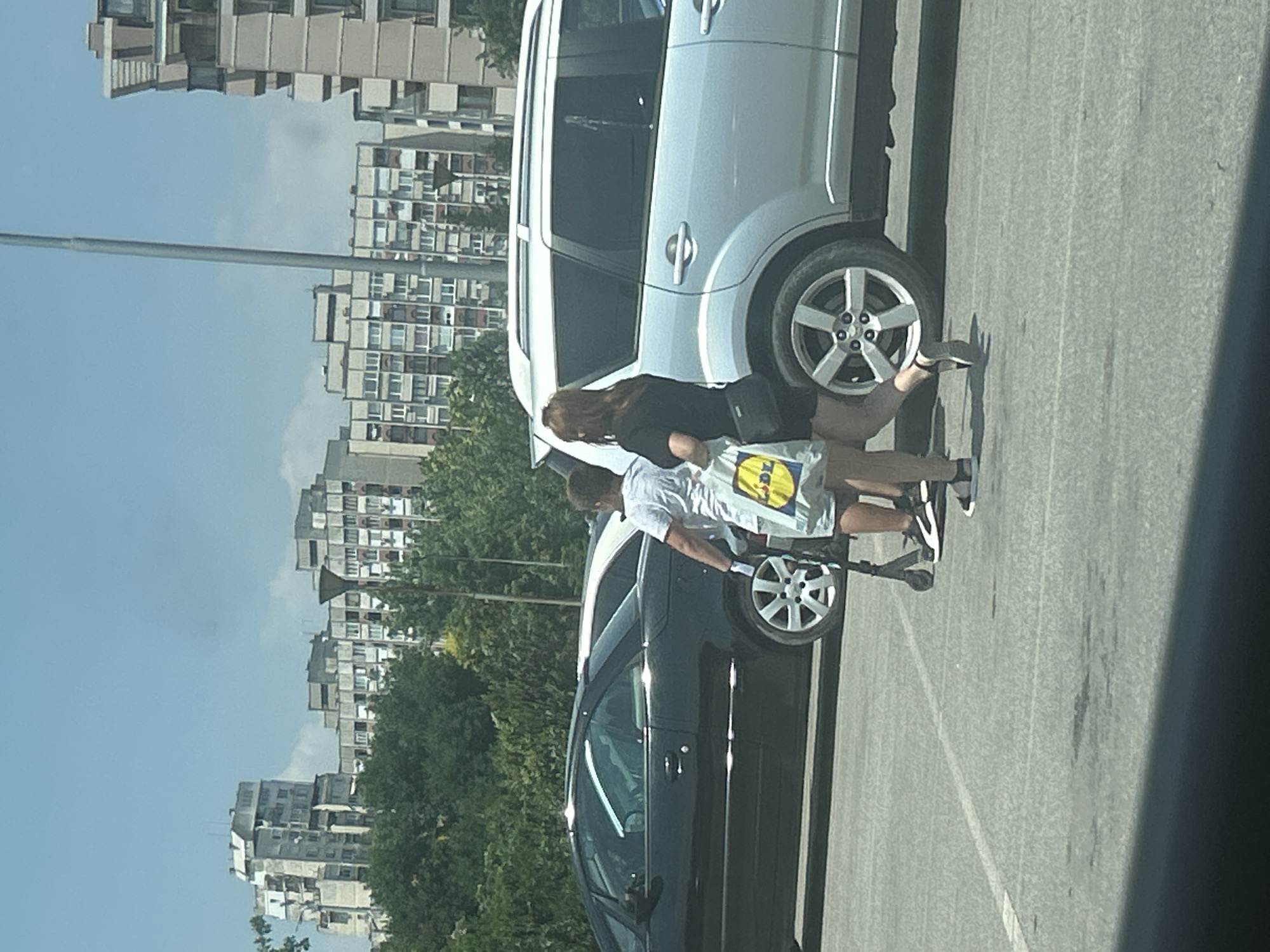 In Plovdiv, Bulgaria, on a bright, sunny day, a young boy and a young woman are captured in a parking lot. The boy, wearing a white t-shirt and shorts, stands near a scooter, holding what appears to be a white grocery bag with a yellow logo, possibly from Lidl. He is also wearing white gloves. The young woman, dressed in a black t-shirt and shorts, walks beside him, carrying a similar grocery bag.
In the background, two cars are parked: a black sedan to the left and a silver SUV to the right. The SUV's rear passenger-side wheel is visible. Beyond the parking lot, a dense line of trees separates the area from a cluster of tall, residential buildings, characteristic of Soviet-era architecture, with visible balconies and windows. The sky is clear with a few scattered clouds. The overall scene depicts a casual moment in a Plovdiv neighborhood.