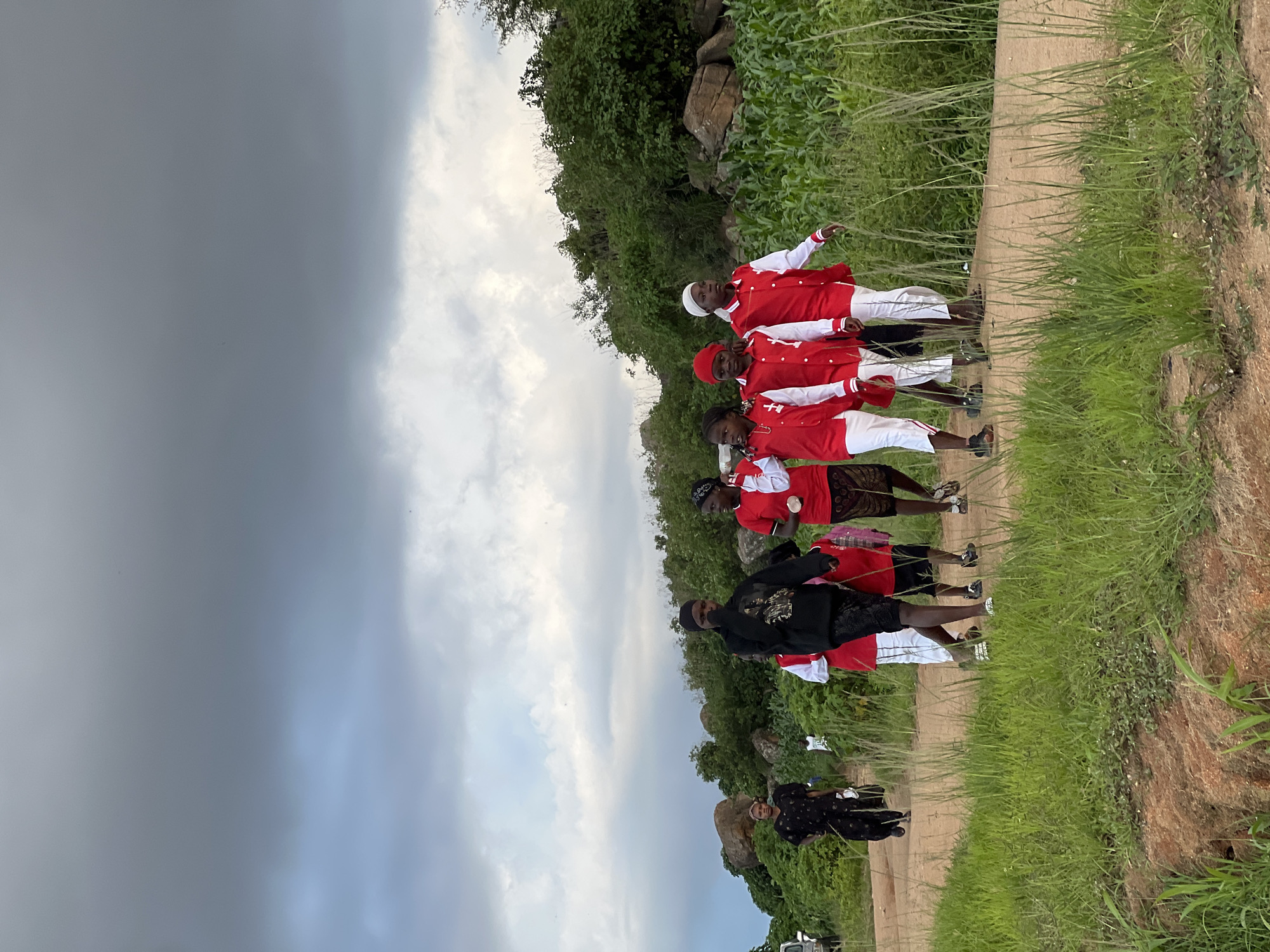 In an outdoor scene, a group of people walk along a dirt road. The sky above is overcast with dramatic clouds. On the left, a woman in a black and white patterned dress walks away from the camera. To her right, a group of children, mostly in red and white uniforms, walk towards the camera. One of the children is in a black hoodie. In the background, lush green vegetation and rocky outcrops are visible, with a cornfield to the right. The overall setting appears to be rural, possibly on the outskirts of Jos, Nigeria, given the vegetation and terrain.