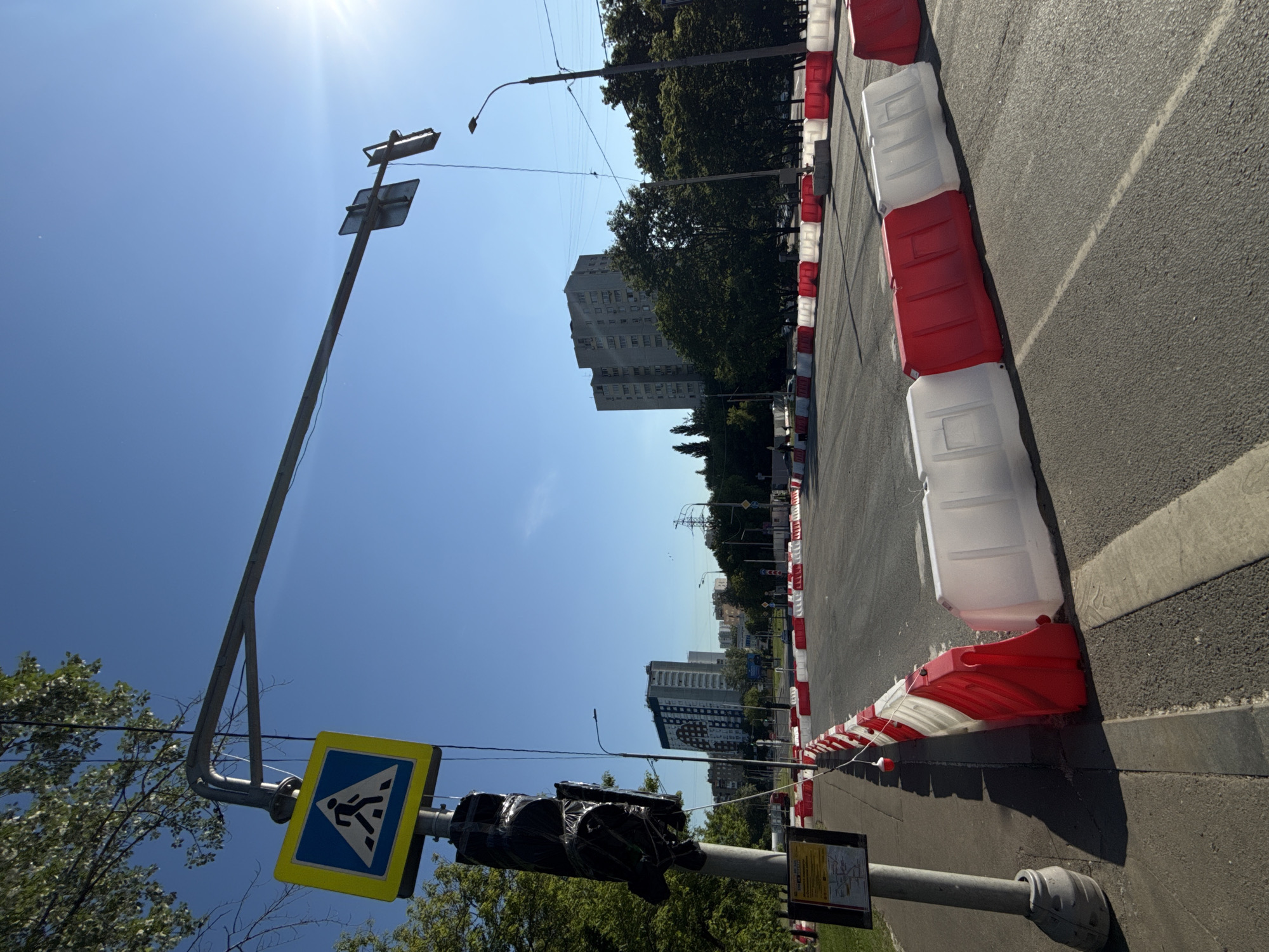 This photograph captures a bright, sunny daytime street scene in Moscow, Russia, rotated 90 degrees clockwise from its natural upright orientation.
The image is dominated by a clear blue sky with the sun visible as a bright flare in the upper left corner (when viewed upright). A tall, light-colored metal pole stands prominently on the left side of the frame, extending towards the sky. Attached to this pole is a blue and yellow pedestrian crossing sign, featuring a white triangle with a black walking figure. Below it, a traffic light is visible, completely wrapped in black plastic or a bag, indicating it is out of service or undergoing maintenance. Further down the pole, near the bottom of the frame, there is another sign with what appears to be a bus schedule or route information, though the text is illegible.
To the right, a paved road surface with a single white line runs alongside a sidewalk. A series of red and white plastic barriers, typically used for road work or traffic control, line the edge of the road, extending into the distance. These barriers are arranged alternately in red and white sections, creating a distinctive pattern.
In the mid-ground and background, various multi-story residential or commercial buildings are visible amongst green trees, suggesting an urban environment. Overhead power lines or utility cables crisscross the sky. The overall impression is one of an active urban setting where some form of road infrastructure work is taking place under clear weather conditions.
