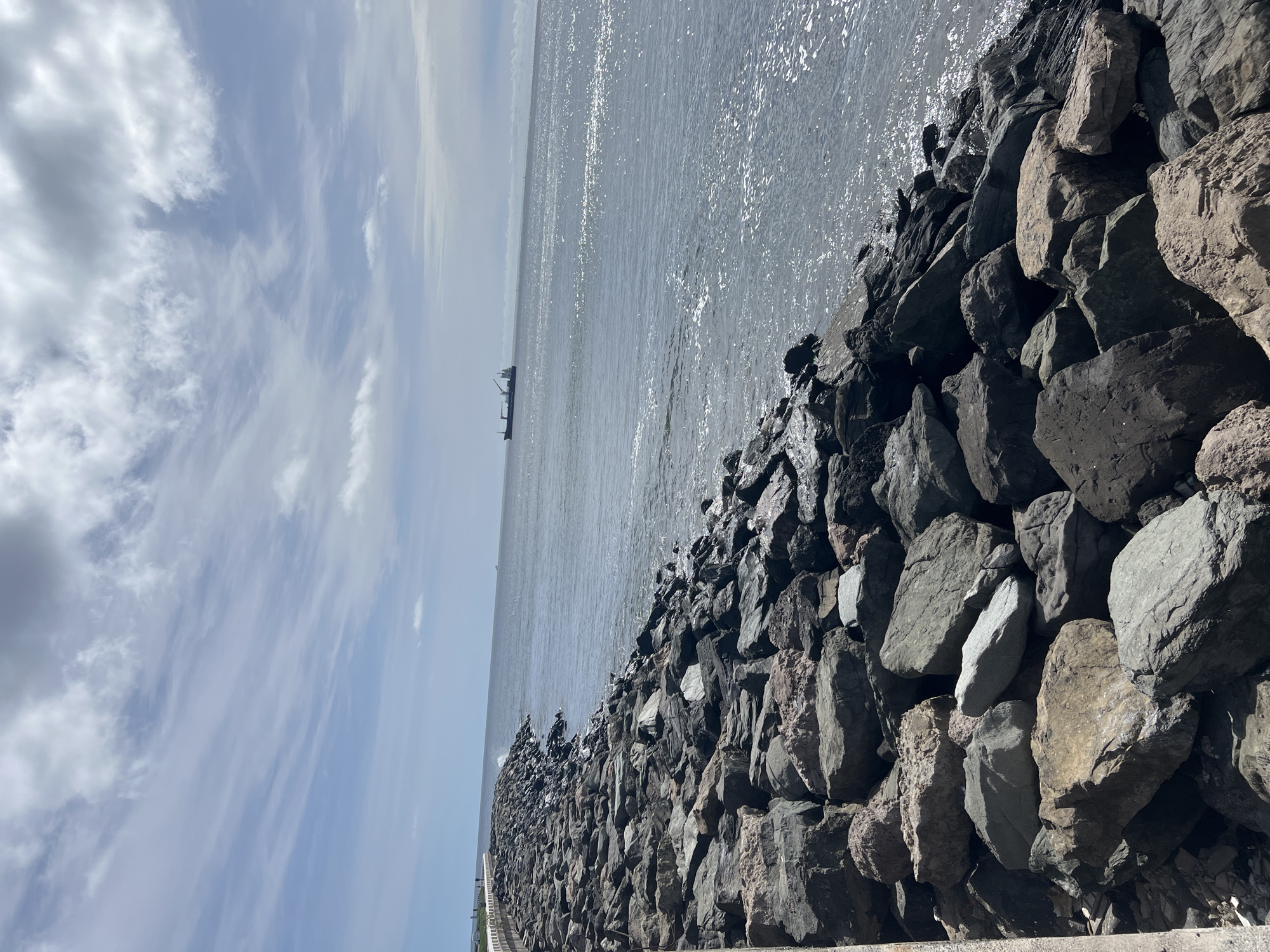 The image shows a coastal scene in Las Palmas, Spain. In the foreground, a breakwater or barrier is constructed from large, irregularly shaped rocks. The rocks are various shades of gray and brown, with some appearing darker, possibly from being wet. Beyond the rocks, the Atlantic Ocean stretches out to the horizon, its surface reflecting sunlight and appearing shimmering. In the distance, a cargo ship is visible. The sky is pale blue with scattered clouds. The image is taken at an angle.