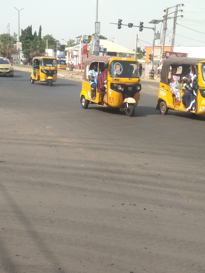 The image shows a street scene in New Karu, Nigeria, at the intersection of 8.998 latitude and 7.571 longitude. Three yellow, three-wheeled vehicles are parked in the middle of the street. The three vehicles are known locally as "Keke Napep" and are frequently used as a form of public transportation. Two of the Keke Napep vehicles are occupied by passengers. In the first, a man is seated in the driver's seat, and a woman is seated in the passenger seat. In the second, three women are seated in the rear, with an obscured driver seated at the wheel. The third vehicle is unoccupied. All three vehicles are facing forward. In the background, one car is visible, facing forward. The road is asphalt. The day is sunny. Power lines, a traffic light, and buildings are visible in the background.