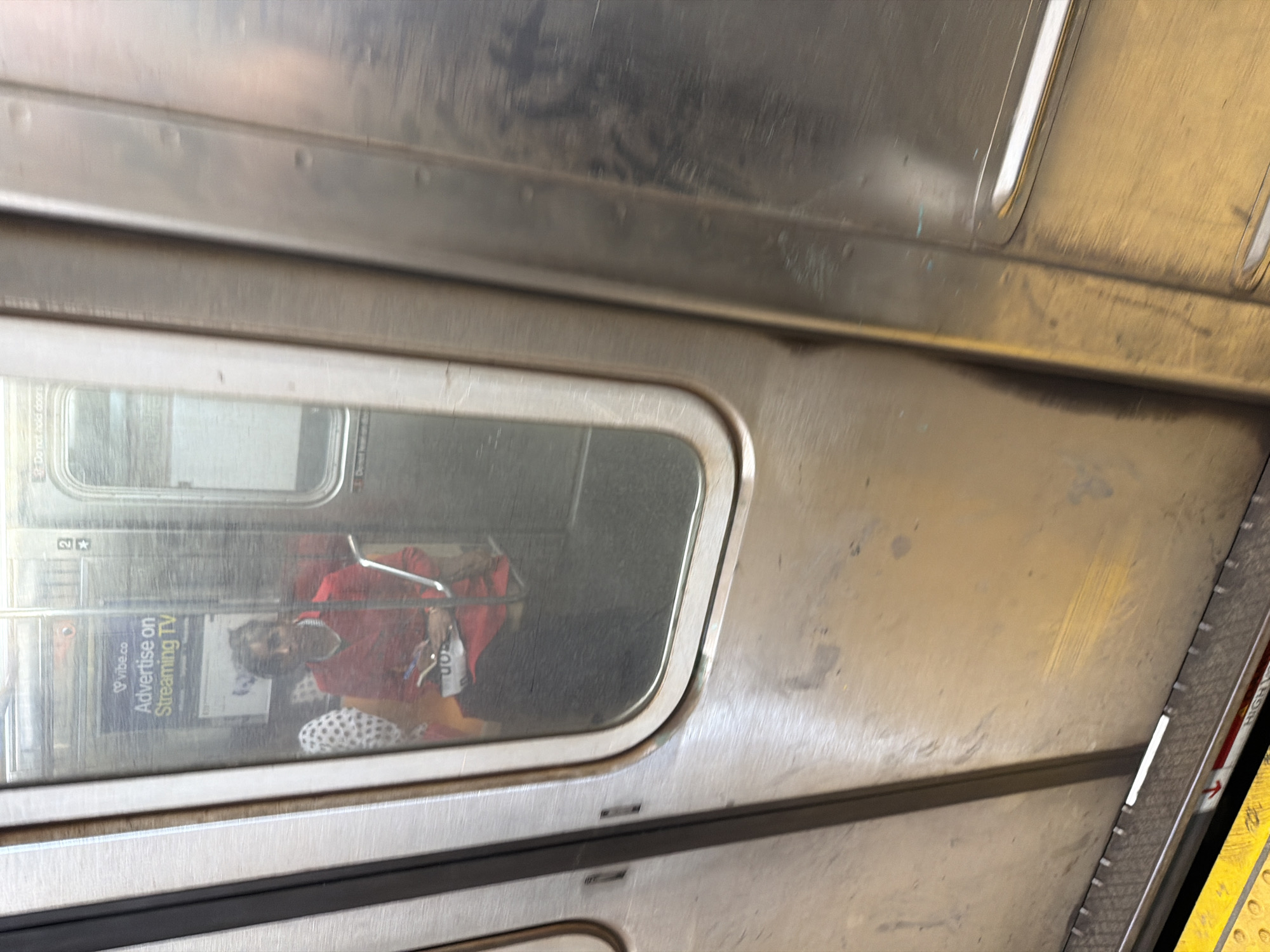 The image shows the interior of a subway car, likely in New York City, United States. A window reflects the interior of another car, including a seated person wearing a red shirt and patterned pants. A "vibe.co Advertise on Streaming TV" advertisement is visible on the wall of the car reflected in the window. The subway car's interior is predominantly metallic, showing signs of wear and age. A yellow safety line can be seen on the right of the frame.