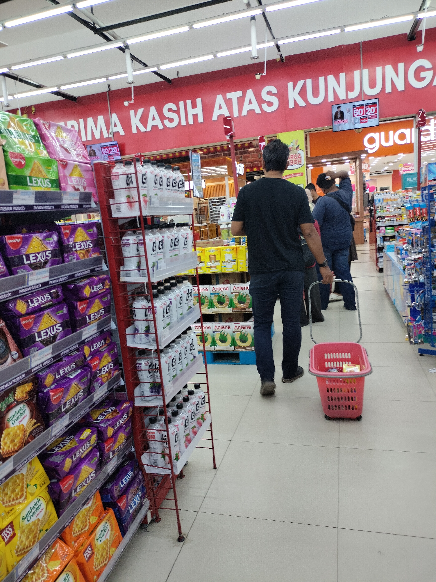A person, likely male, wearing a black t-shirt and dark pants, walks through a grocery store with a red shopping basket in hand. The basket appears to contain a few items. In the background, other shoppers are visible, and shelves are stocked with various products, including snacks and beverages. A prominent red banner with white text, "PRIMA KASIH ATAS KUNJUNG," is displayed overhead. A digital screen shows "50 20." The floor is tiled, and fluorescent lights illuminate the store. The scene is consistent with a typical supermarket environment.