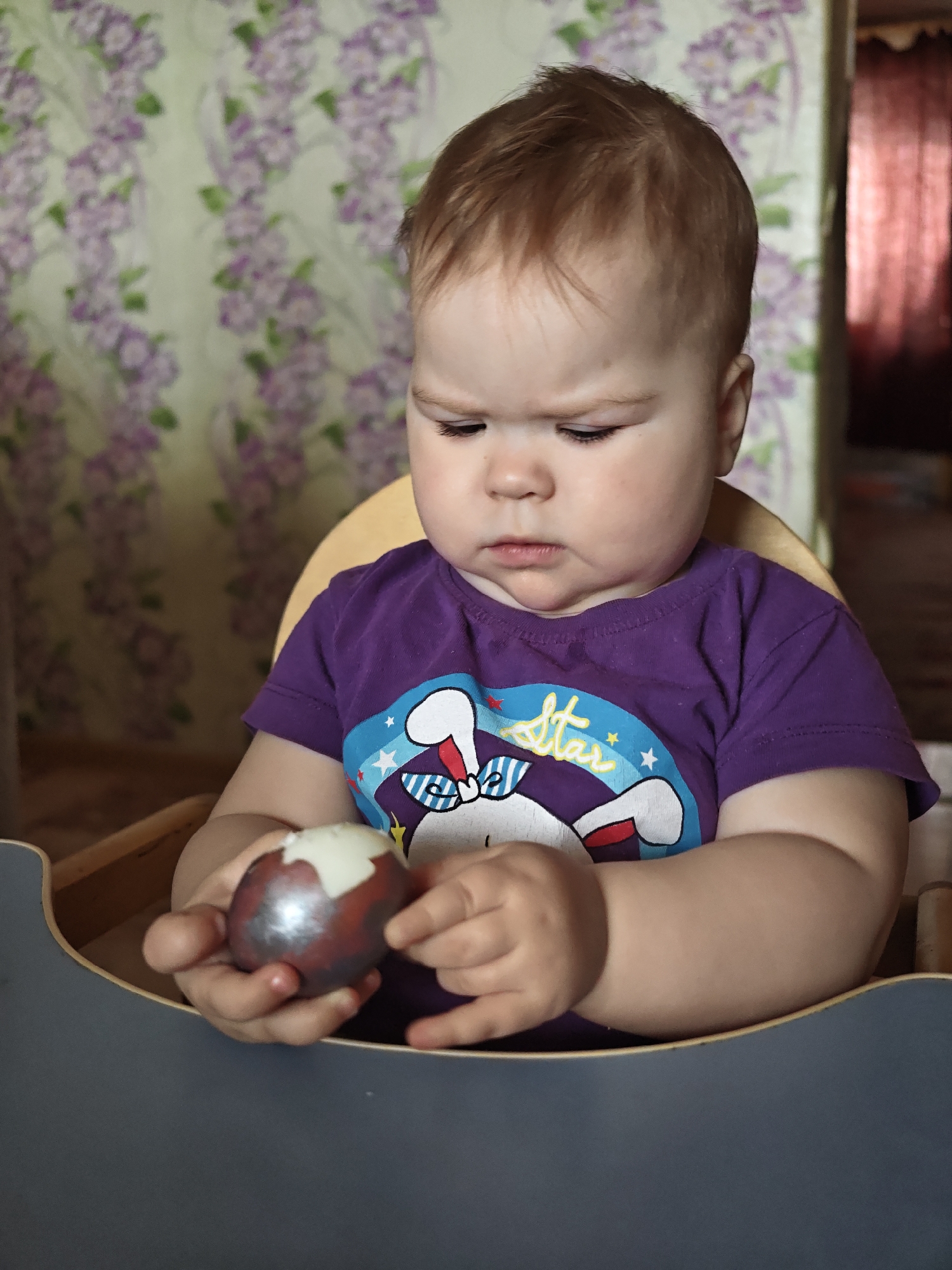 A baby sits in a blue high chair with a yellow seat. The baby wears a purple t-shirt with a rabbit design. The baby holds a partially peeled egg. The background shows a wall with a purple floral pattern and a glimpse of a dark curtained area. It is not possible to verify the location as Уральское, Russia from the image alone.