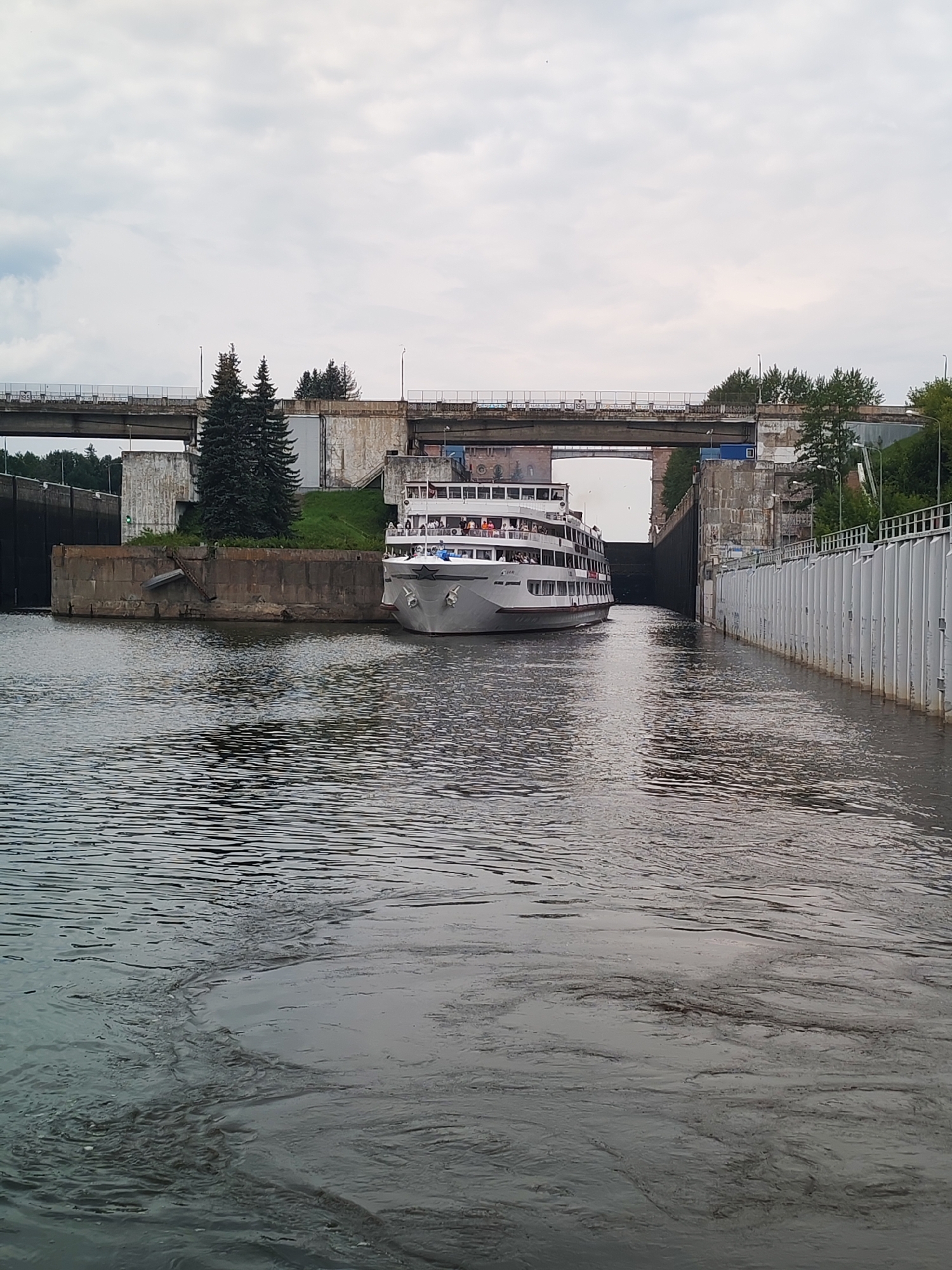 In Bol'shie Mkhi, Russia, a white cruise ship is navigating a concrete canal. The ship has multiple decks with visible passengers on the upper levels. To the right of the canal, a white vertical structure lines the bank, likely part of the lock system. To the left, the canal opens up, with a concrete embankment and a grassy slope leading up to a bridge structure. Two tall, dark green evergreen trees are situated on this embankment. The sky above is overcast with gray clouds.