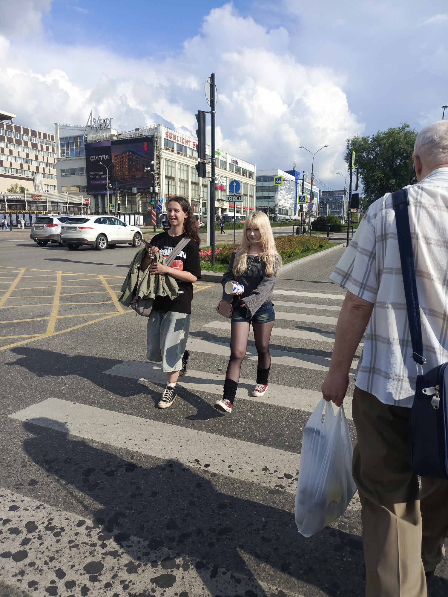 An outdoor, medium shot captures three individuals crossing a pedestrian crosswalk on a sunny day in the city of Perm, Russia. The foreground features the back of an older man wearing a plaid shirt and khaki pants, carrying a white plastic bag in his right hand and a navy blue satchel on his right shoulder. To his left, two younger women are walking across the crosswalk. The woman on the left, with long brown hair, wears a black t-shirt with a graphic print, light blue denim culottes, and black and white Converse sneakers. She carries a green jacket slung over her left shoulder and a gray bag in her right hand. The woman on the right, with blonde hair, wears a grey long-sleeved shirt, denim shorts, black tights, and red and white sneakers. She holds a white cup in her right hand and a pink purse on her left shoulder. Behind the pedestrians, white cars are visible on the street, and buildings line the background, including a building with signage that appears to read "СИТИ" and "SUNLIGHT". The sky is a mix of blue with scattered white clouds. The ground is an asphalt crosswalk marked with white stripes.