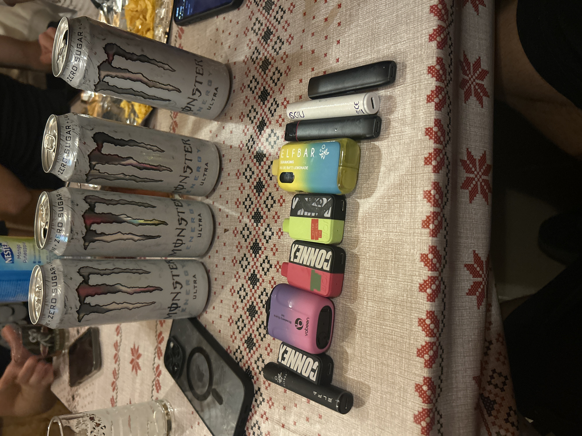 A top-down medium shot captures a table covered with a patterned tablecloth. Four Monster Energy Ultra zero-sugar cans stand upright on the table. To the left of the cans, a clear glass with condensation is visible, and a smartphone is also present on the table. In the foreground, a row of vape pens is laid out horizontally. The vape pens vary in color and branding, including Elfa, Connex, Vabeen, and Elfbar. Some of the vape pens have visible text indicating flavors or brand names. The tablecloth features a repeating pattern of red floral motifs and geometric designs. The background is blurred, showing glimpses of people's hands and arms, suggesting a social gathering. The lighting is ambient, casting soft shadows.