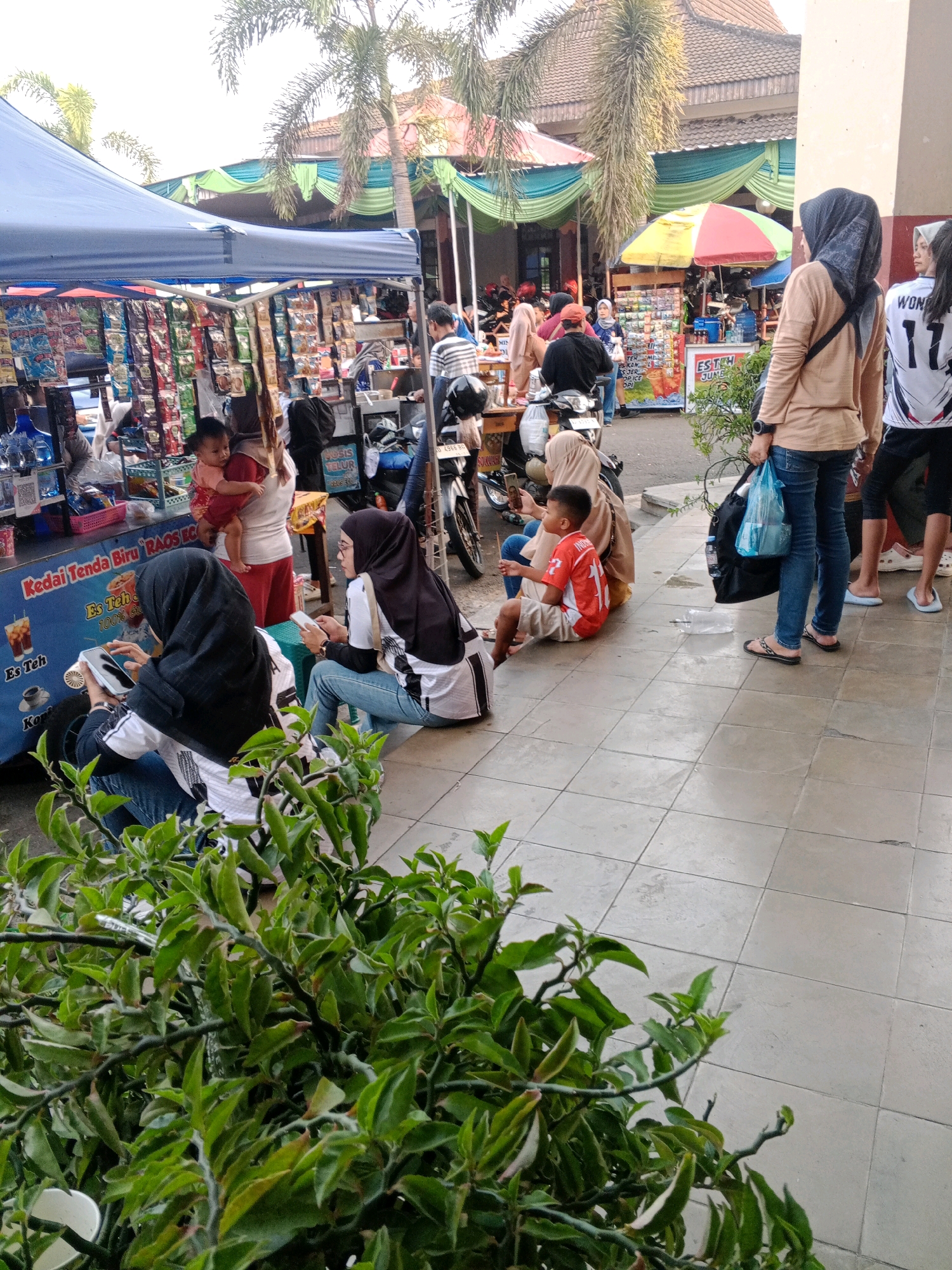 The image depicts a busy street scene, likely a market or roadside vendors, in Wuryorejo, Indonesia. In the foreground, a woman wearing a black hijab and a white and black patterned long-sleeved shirt sits on the ground, holding a white mobile phone. To her right, another woman, also in a hijab and similar attire, sits on a green stool, looking at her mobile phone. Behind them, a young boy in an orange shirt and shorts sits on the tiled ground, his gaze directed downwards. A woman in a beige top and blue jeans stands to the right, holding a large black bag and a blue plastic bag. Several stalls are visible in the background, selling various items, including packaged goods and drinks. Palm trees and awnings provide shade. A motorcycle is parked near one of the stalls. The ground is paved with light-colored tiles.