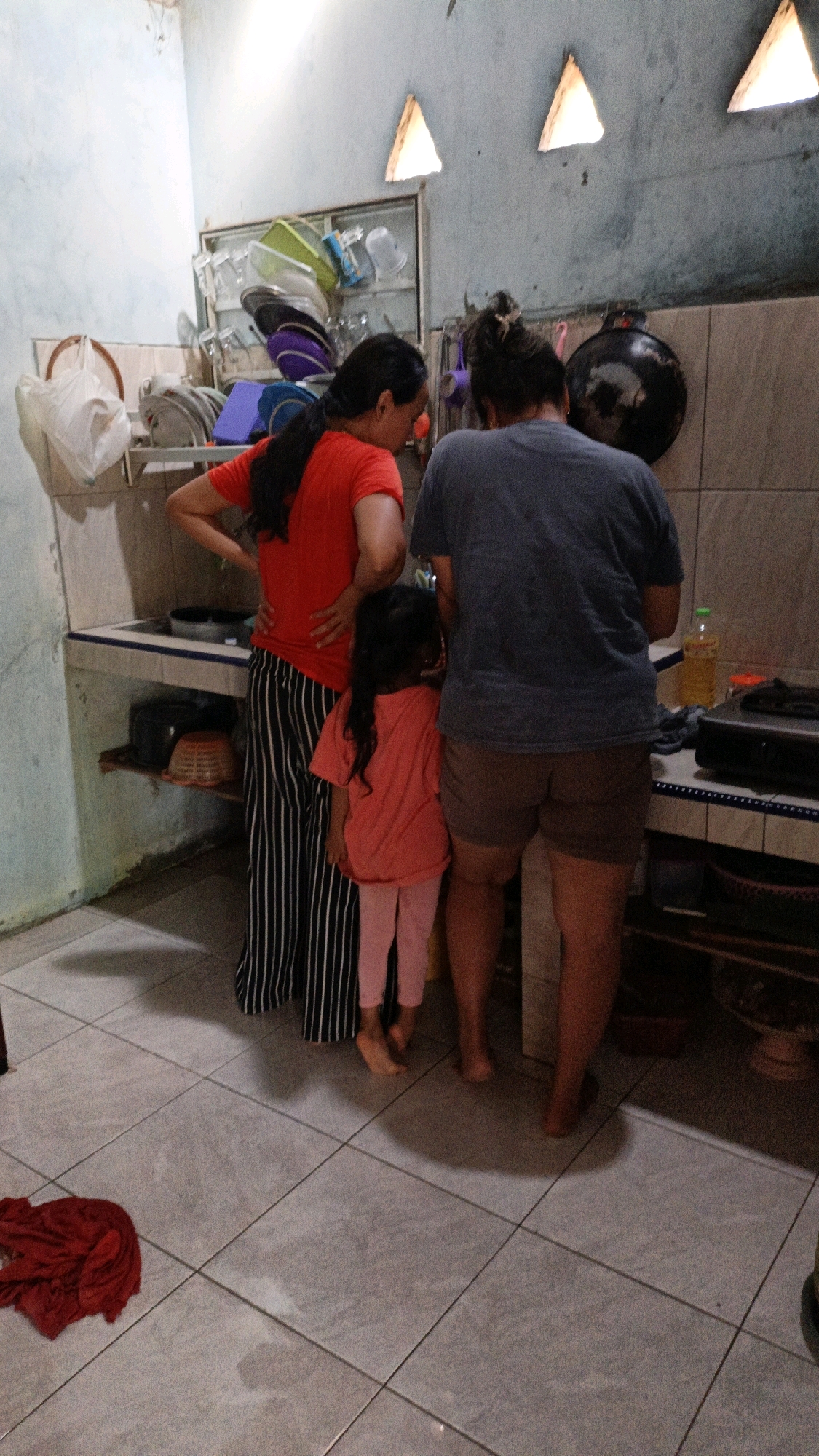 The image depicts three individuals, two adults and one child, standing in what appears to be a kitchen area in a residential setting in Suwawal Timur, Indonesia. The lighting in the room is dim, with natural light entering from triangular openings in the upper wall. The walls are tiled with a light-colored material, and the floor is covered with square tiles.
On the left, an adult with dark hair pulled back is wearing a red t-shirt and black and white striped pants. Their right hand is on their hip, and their left arm is bent at the elbow. To their right, a young child with dark, shoulder-length hair is wearing a coral-colored t-shirt and light pink leggings. The child is facing away from the camera, looking towards the center of the scene. On the right, another adult, also facing away from the camera, is wearing a dark grey t-shirt and brown shorts.
The background reveals kitchen elements. A shelf unit with various items, including bowls and what appear to be containers, is visible. A white bag is hanging on the wall to the left. A black pan is hanging on the tiled wall to the right. A stovetop is present, and a bottle of yellow liquid, possibly cooking oil, is on the counter. There is also a dark object on the counter to the right of the stovetop. A red cloth is lying on the floor in the lower left corner of the image. The overall impression is of a utilitarian, domestic space.