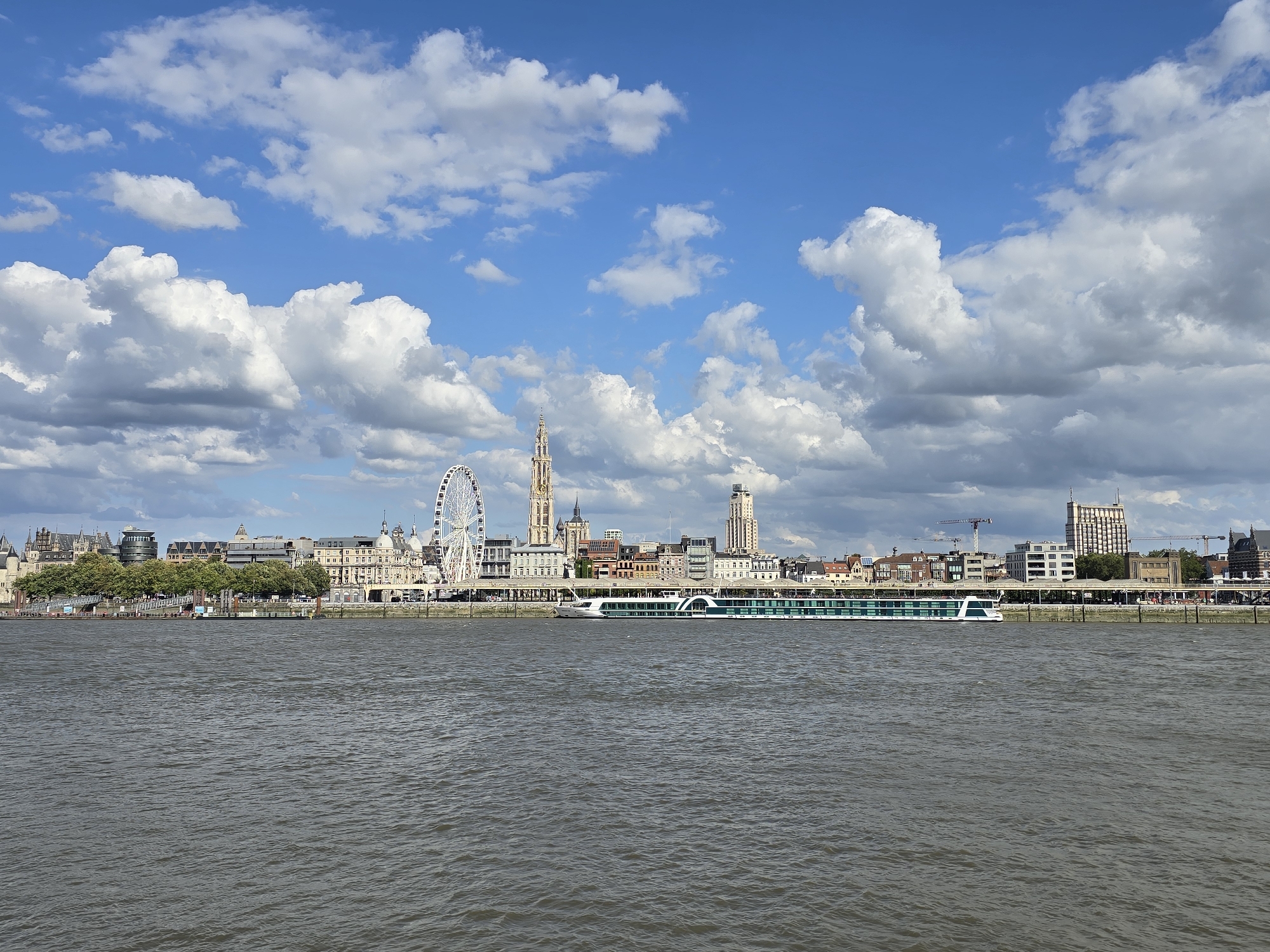 The image captures a wide-angle view of the Antwerp skyline along the Scheldt River on a partly cloudy day. In the foreground, the dark, rippling water of the river dominates the lower half of the frame. The middle ground showcases a long, modern white and green river cruise ship docked near the embankment. Behind the ship, the Antwerp cityscape unfolds with a mix of historical and contemporary architecture. Prominent landmarks include a large Ferris wheel on the left, the tall spire of the Cathedral of Our Lady, and several other multi-story buildings, including a distinctive tall, modern building on the right. A line of trees separates the riverfront from the buildings. The background is filled with a bright blue sky adorned with voluminous white and gray cumulus clouds. The overall scene depicts a vibrant urban waterfront with a clear view of Antwerp's iconic structures.
