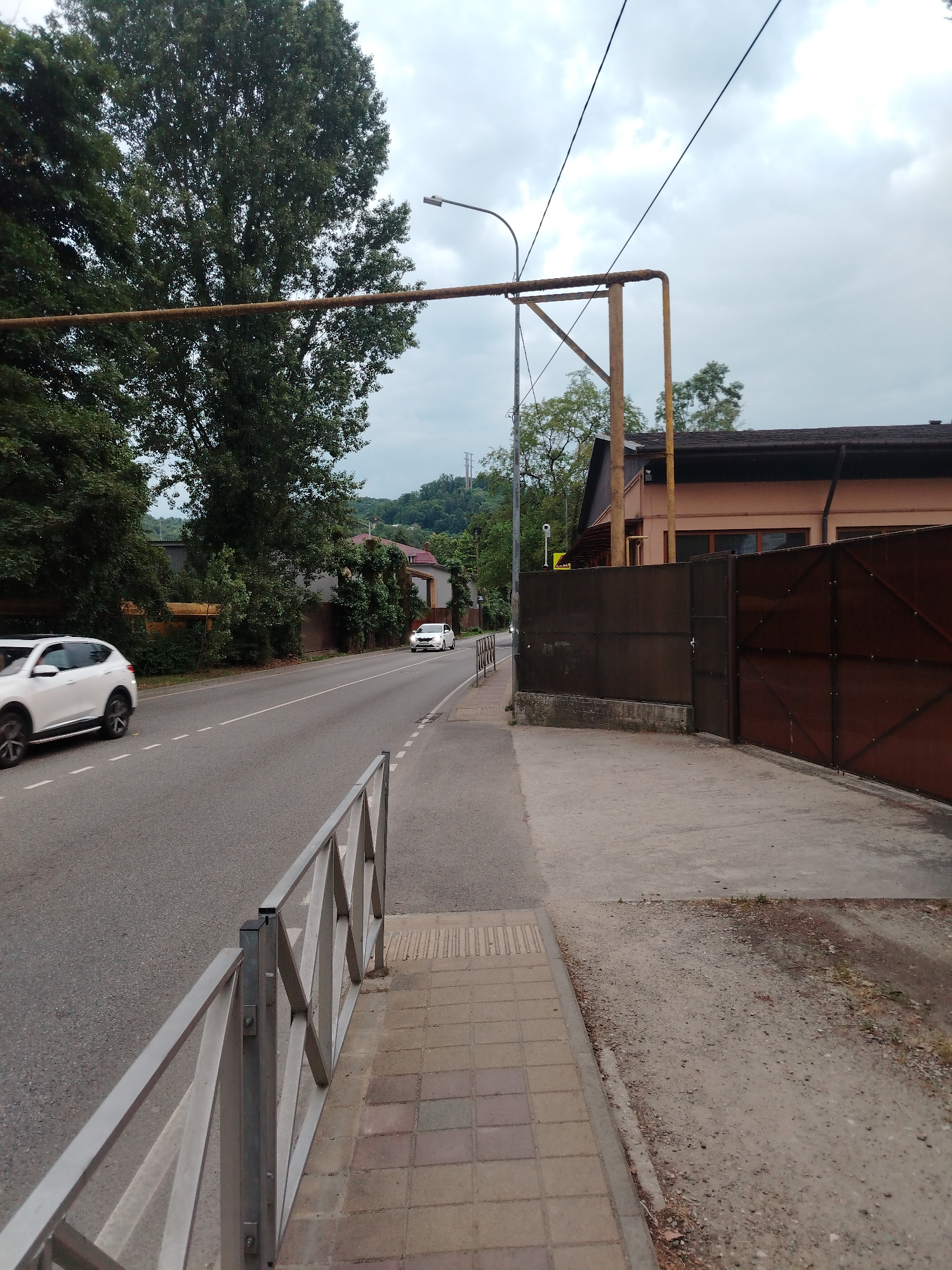 A street scene in Sochi, Russia, features a two-lane asphalt road with a white car in the left lane and another white car in the right lane heading away from the camera. To the left of the road, a metal railing separates a paved sidewalk from the street. Beyond the sidewalk, dense green trees line the area. On the right side of the road, a concrete sidewalk runs alongside a brown metal fence and a light brown building with a dark roof. Overhead, power lines and a rusted pipe structure are visible against a cloudy sky. In the background, hills covered in trees rise under the overcast sky.