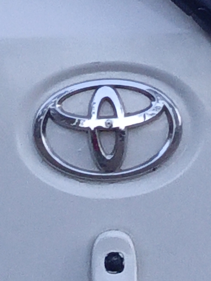 The image shows a close-up of a Toyota emblem. The emblem is silver and circular, with three overlapping oval shapes. It is mounted on a white surface, likely a vehicle. Below the emblem, there's a small, white, rectangular shape with a black indentation. The surface around the emblem is slightly textured.