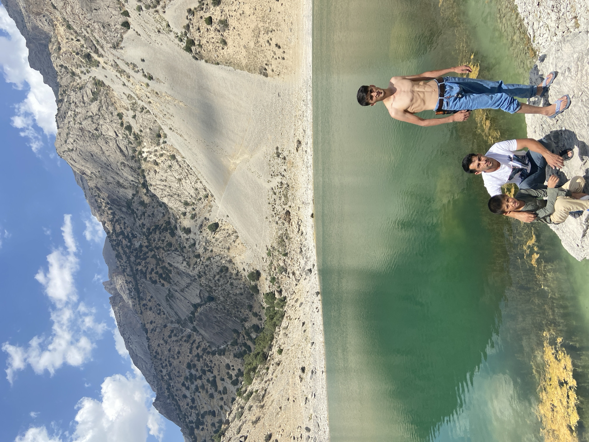 In the city of Ghazza, Tajikistan, a scenic mountainous landscape surrounds a pristine, emerald-green lake. The water's surface is calm, reflecting the azure sky dotted with white clouds. The steep slopes of the mountains are covered with rugged terrain, sparsely vegetated with small shrubs and trees.

In the foreground, three individuals are positioned near the lake's edge. A young boy, dressed in a long-sleeved olive shirt and light-colored pants, sits beside a man wearing a white t-shirt and dark pants. The man has his arm around the boy's shoulders, suggesting a familial connection. To their right, a young man stands shirtless, wearing blue jeans and flip-flops. He appears to be posing for the photograph, with a lean physique.

The ground around them is rocky and appears to be a natural shoreline. The overall scene conveys a sense of natural beauty and outdoor recreation.