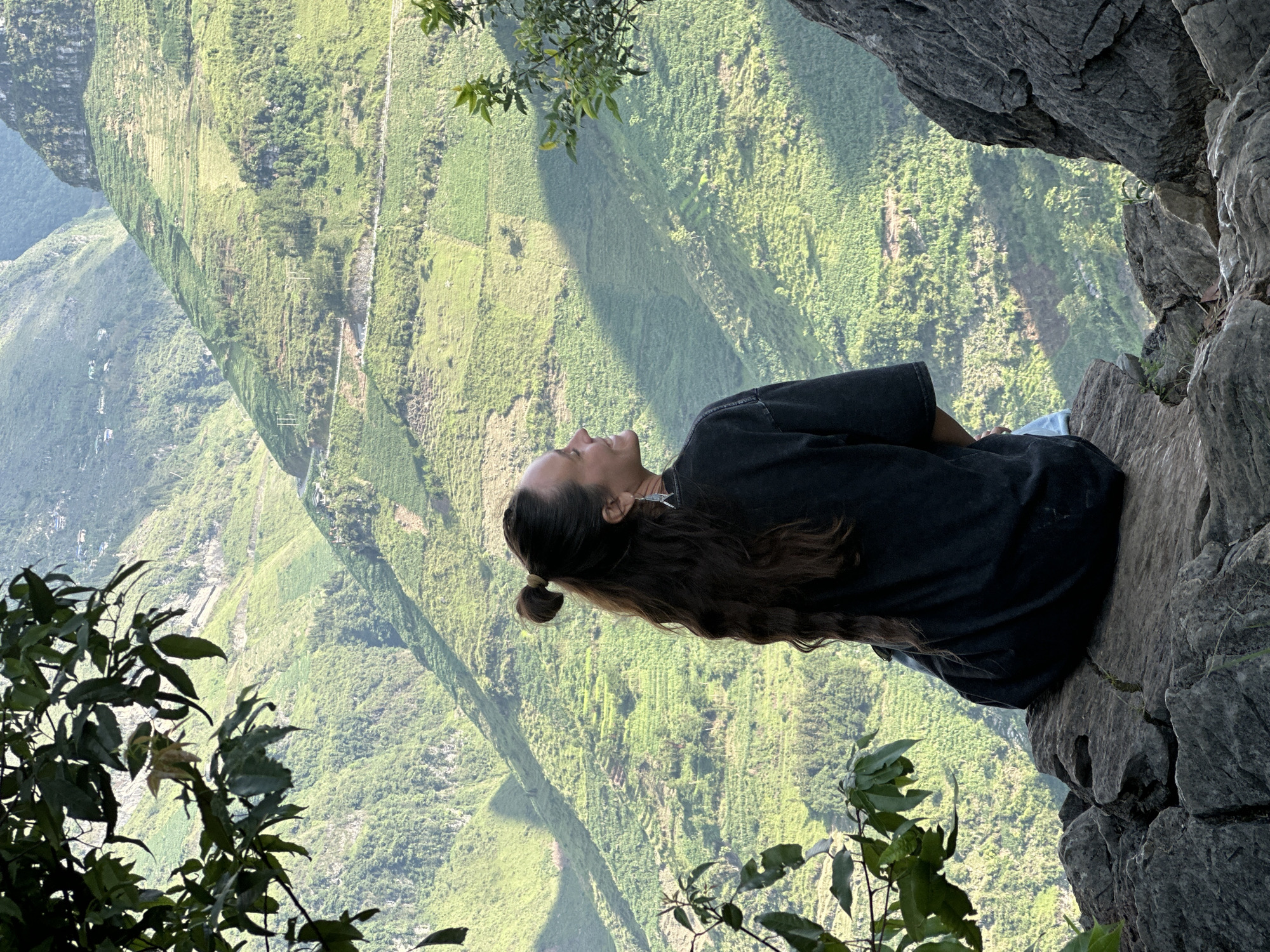 A woman is sitting on a rocky outcropping. The woman is facing left, with her head tilted back. She has long brown hair pulled back in a ponytail. She wears a dark t-shirt and light-colored pants. The background is a steep, green landscape with dense vegetation. There are leaves and branches in the foreground. The location is in Đồng Văn Commune, Vietnam.