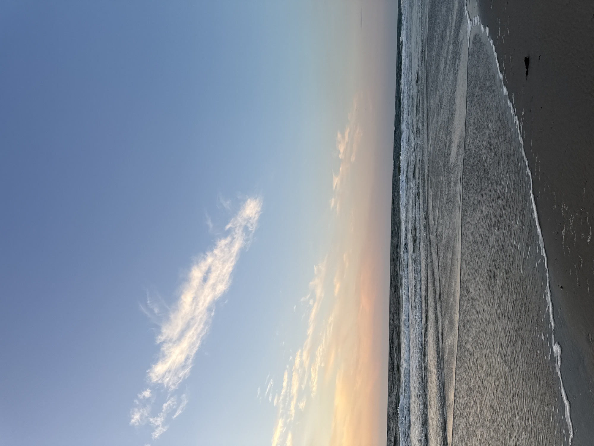 The image depicts a serene beach scene at sunset on the island of Langeoog, Germany. The sky transitions from a clear blue at the top to soft hues of peach and pink near the horizon, where wispy clouds are scattered. The ocean is calm, with gentle waves rolling towards the sandy shore. The foreground shows wet sand, reflecting the sky's colors, and a thin line of white foam from the receding tide. The overall atmosphere is tranquil and peaceful, characteristic of a sunset on the North Sea coast. No people or specific landmarks are visible in the image, focusing entirely on the natural elements of the seascape.