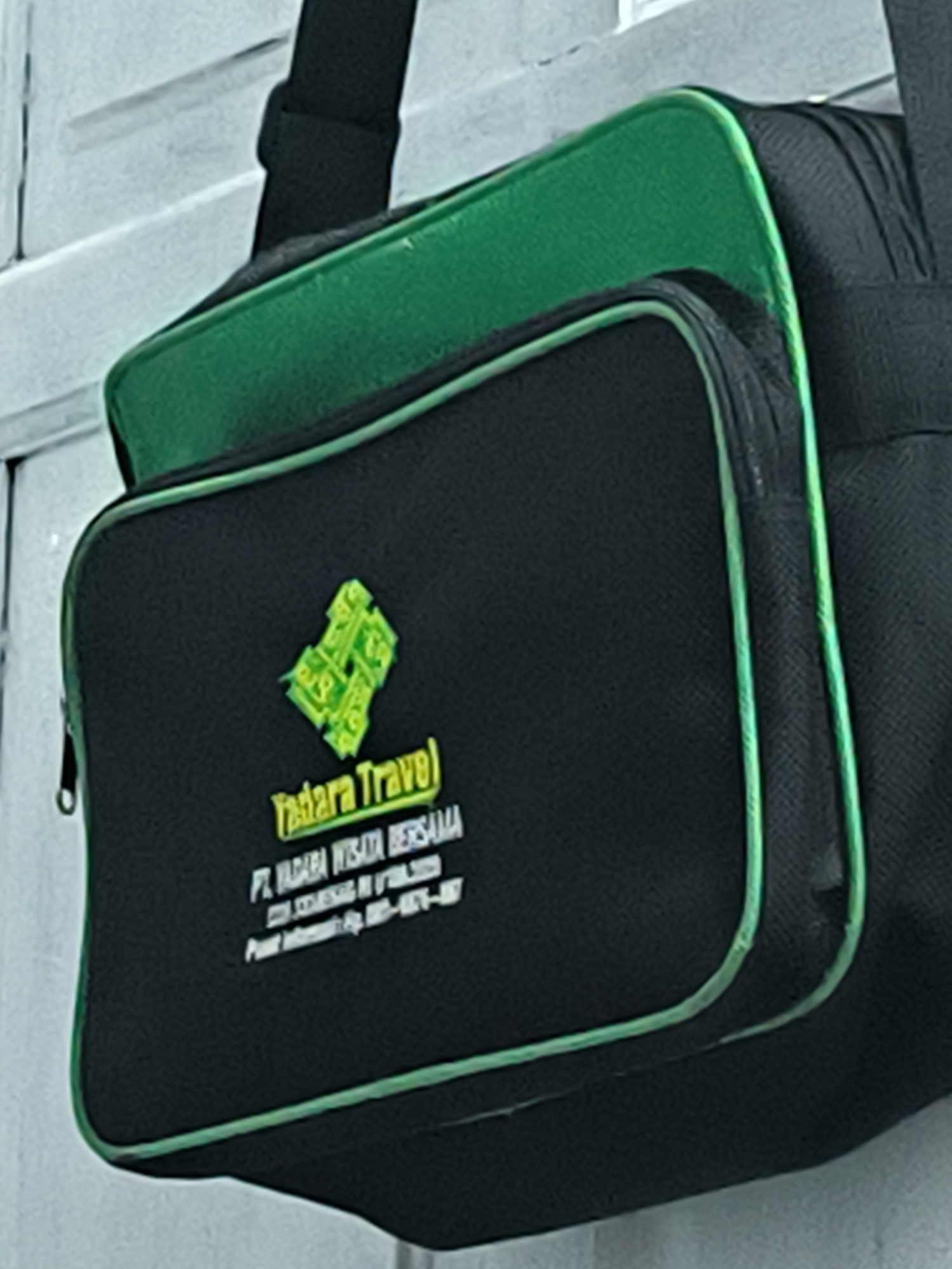 A black shoulder bag with vibrant green accents hangs against a blurry, light-colored background, possibly a wall or fence with horizontal lines. The bag features a main compartment and a smaller front zippered pocket, both edged with bright green piping. A black shoulder strap is visible at the top.
The front pocket of the bag displays a prominent green and yellow logo, which appears to be a stylized design, possibly depicting abstract shapes or elements resembling currency. Below the logo, the text "Tadara Travel" is printed in a bold, yellow-green font. Further text, in a smaller, lighter font, identifies the company as "PT. TADARA WISATA BERSAMA". An address is also visible: "JL. COTGURENA KEC. KUTAMAKMVR", followed by a contact number: "HP. 0823-8717-1077".
There are no people or specific activities depicted. The scene is static, focusing solely on the branded bag. The lighting suggests ambient daylight, but the time of day and weather conditions are not clear. The image provides a close-up view of the bag's branding, indicating its use by a travel company based in Lhokseumawe, Indonesia.