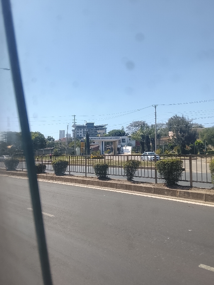 A view from a vehicle, likely a bus, shows a road with a metal railing separating it from a grassy area. Small, manicured bushes line the railing. Beyond the railing, a building with signage reading "KENYA INSTITUTE OF CURRICULUM DEVELOPMENT" is visible. A white car is parked nearby. In the background, a modern building with multiple stories and a flat roof stands amidst trees. Utility poles with wires cross the sky. The sky is a clear, bright blue.