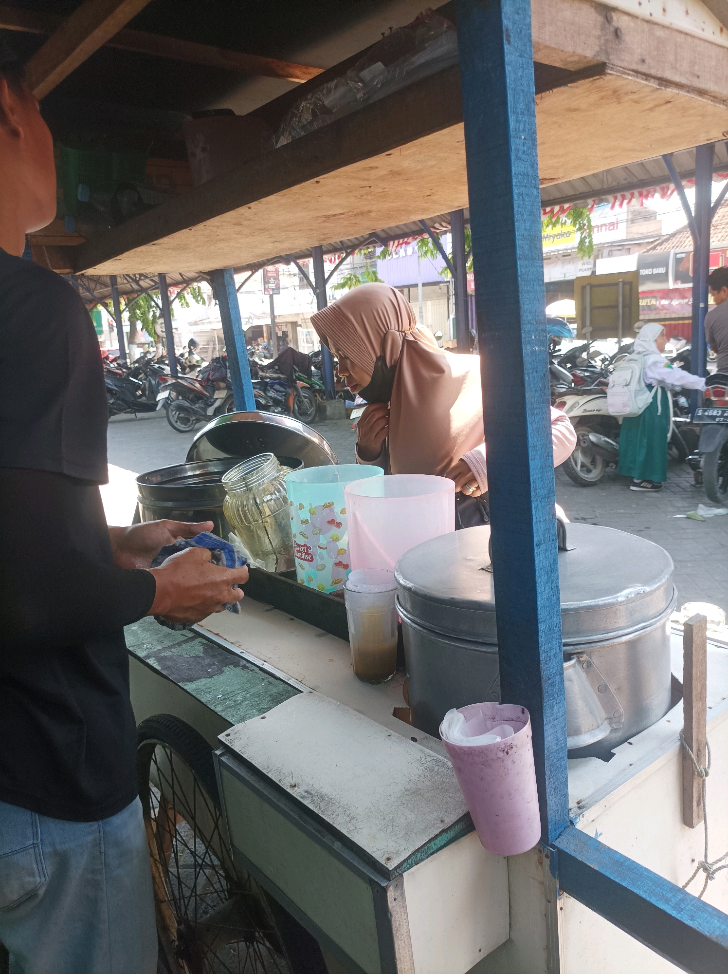 The image shows a street scene in Sumuragung, Indonesia, with a food cart as the central focus. A person wearing a black t-shirt and blue jeans stands on the left side of the frame, facing away from the viewer, and is holding a blue cloth in their hands. To the right of this person, on the food cart, are several objects including a large metal pot with a lid, a glass jar, a light blue patterned cup, and a pink plastic cup containing white straws. Behind the food cart, a woman wearing a beige headscarf is partially visible, looking to the right. In the background, there are motorcycles parked and people walking, suggesting a bustling street environment. The food cart appears to be made of wood and metal, with a blue wooden support beam prominently featured. The overall lighting suggests it is daytime.
