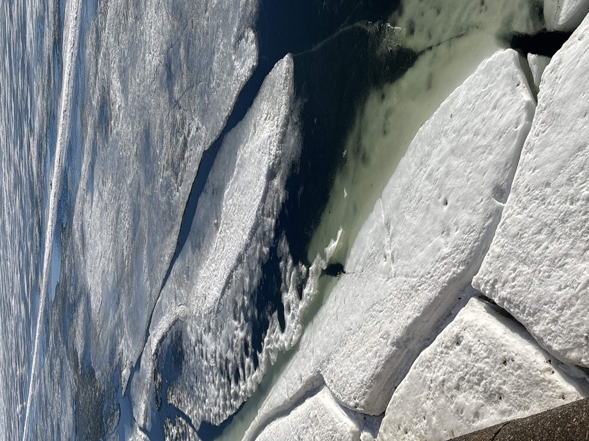 The image depicts a body of water, possibly a river or lake, partially covered with thick layers of ice. The ice is broken into various shapes and sizes, with visible cracks and crevices separating the ice chunks. The ice appears dense and white, some parts showing darker shades in the areas where it adjoins the open water. The water visible between the ice formations appears dark in some sections. The scene is set under daylight conditions, with some glare and reflections suggesting sunlight. The image is taken from an elevated perspective, offering a clear view of the icy landscape. There are no people or animals visible in the frame. The exact location in Chaikovsky, Russia, cannot be determined from this image alone.