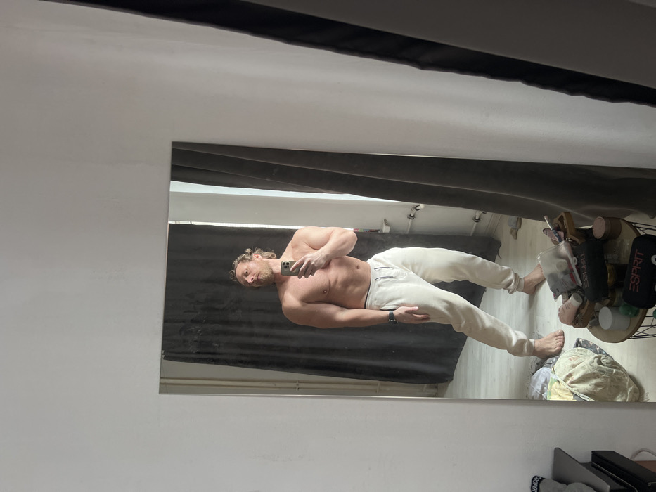 A blond-haired, bearded male individual, shirtless and wearing light cream-colored sweatpants, is depicted in a full-body mirror selfie within an indoor setting in Saint Petersburg, Russia. He stands barefoot, holding a silver-toned smartphone in his right hand to capture the image, with his gaze directed slightly upwards and to his left. A dark smartwatch is visible on his left wrist. The background features white walls, with dark grey curtains partially drawn to reveal a window and diffuse natural light. In the reflection, a radiator or heating pipes are visible along a wall. To the lower right, a small, round, dark-framed table holds various personal care items, including jars, bottles, and a black pouch bearing the "ESPRIT" logo. Additional items, including fabric and stacked objects, are visible on the light-colored floor and a surface to the left.