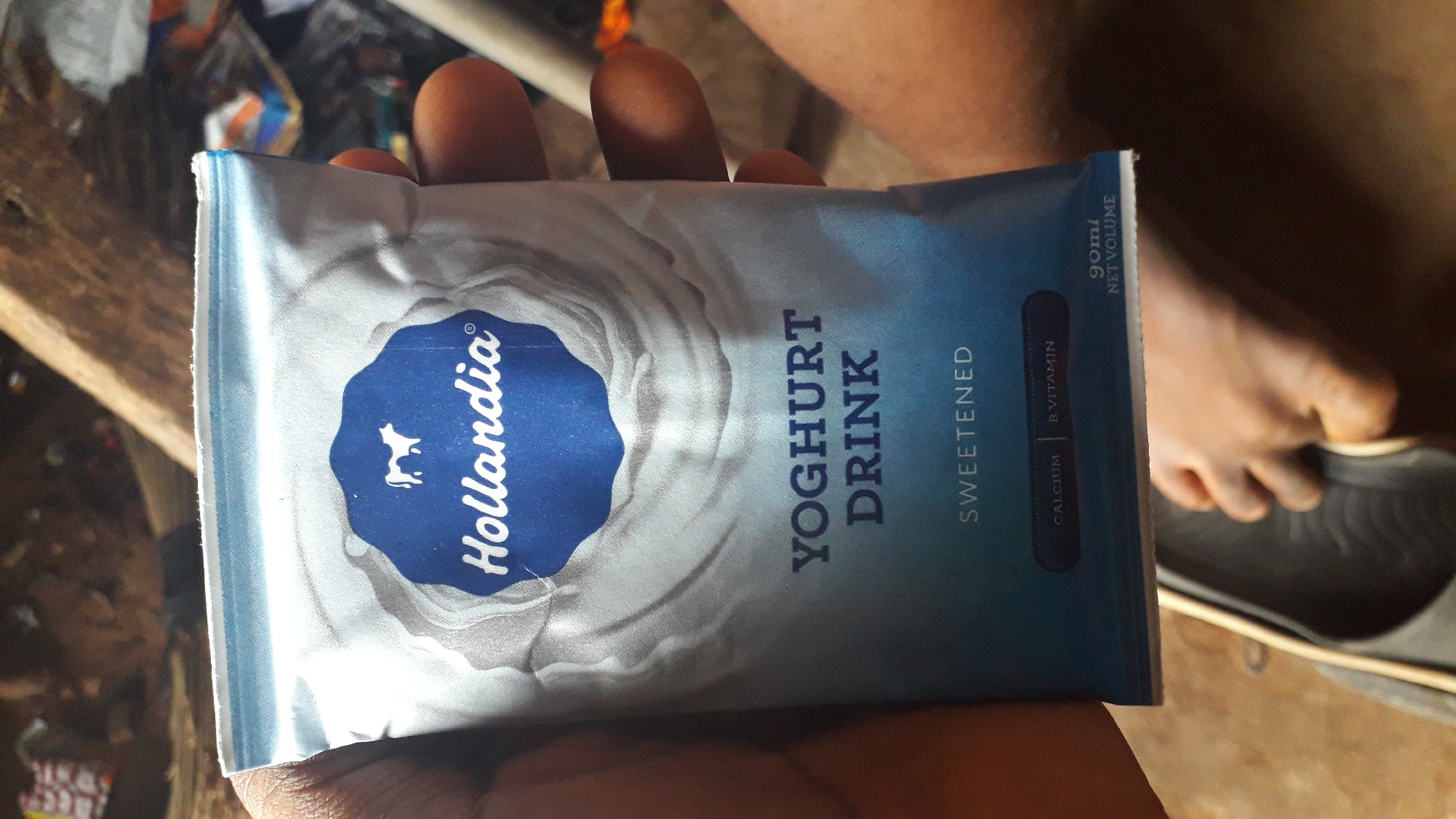 The image shows a person's hand holding a "Hollandia" yogurt drink pouch in what appears to be an indoor setting in Nkpor, Nigeria. The pouch is silver and blue, with the Hollandia logo prominently displayed. The text "YOGHURT DRINK" and "SWEETENED" are visible on the pouch. The background is somewhat cluttered and out of focus, with glimpses of what appears to be miscellaneous items, possible construction material and a dark shoe nearby. The lighting is moderately dim.
