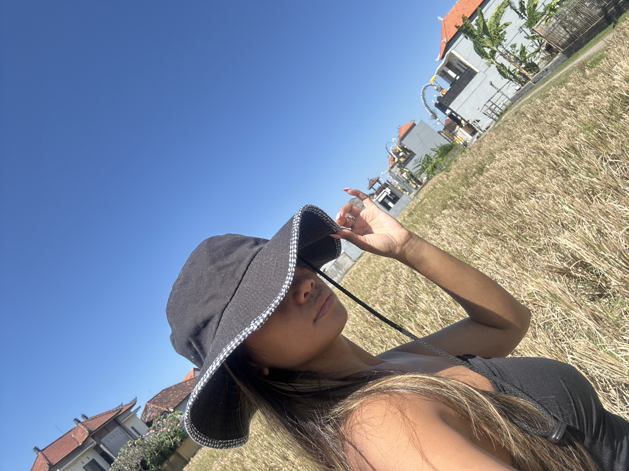 A young woman wearing a black bucket hat with a checkered brim and a black tank top takes a selfie outdoors on a sunny day. Her hand is raised to adjust the hat, shielding her face from the sun. She has long, blonde-streaked brown hair that falls over her shoulders. The background shows a field of dry, golden grass, a few buildings with orange-tiled roofs, and lush green banana trees. The sky is a clear, vibrant blue, indicating a bright and sunny day. The architecture and vegetation suggest a tropical location. The overall mood is relaxed and carefree. No text is visible in the image.