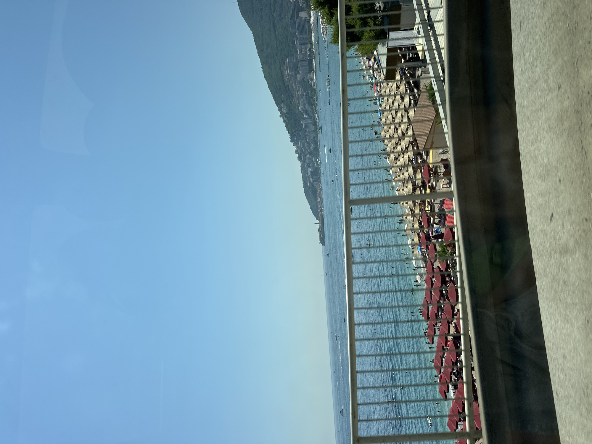 The image shows a view from an elevated position overlooking a bustling beach in Budva, Montenegro. The turquoise water of the Adriatic Sea is calm, with several boats and smaller vessels visible in the distance. Along the coastline, a densely populated beach is filled with numerous beach umbrellas, some in red and others in natural straw colors, providing shade for beachgoers. Many people are seen relaxing on the sand and swimming in the sea.
In the background, the hilly landscape of Budva is dotted with buildings, suggesting an urban environment adjacent to the coast. The sky is a clear, bright blue, indicating a sunny day. A white railing runs across the foreground, partially obscuring the view, which appears to be taken from a vehicle or a walkway above the beach. The overall scene depicts a vibrant summer day at a popular tourist destination.