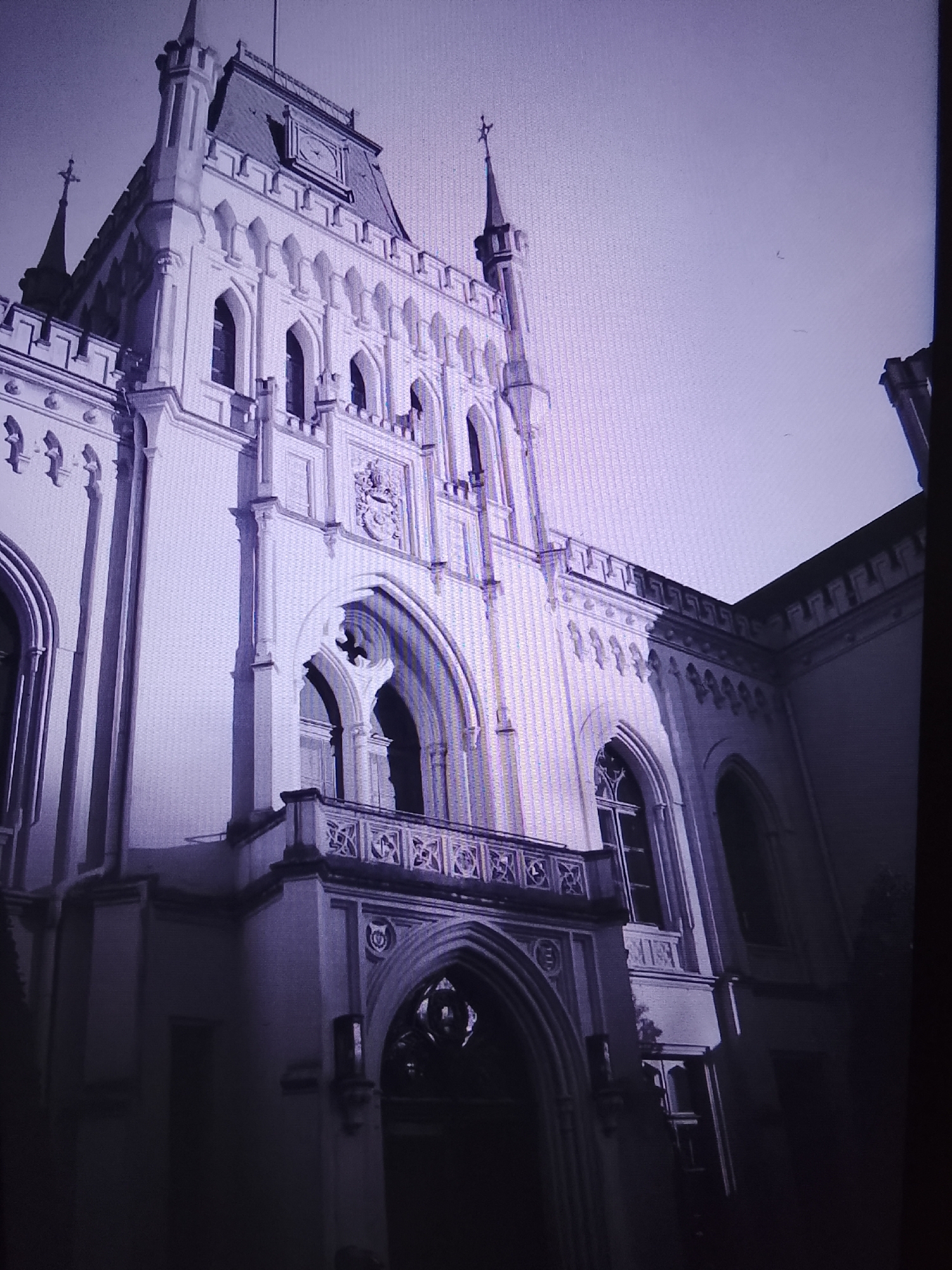 The image shows a large, ornate building with Gothic architectural elements. The building is predominantly white or light-colored, with many arched windows and decorative detailing along its facade. A prominent tower with a spire rises from the left side of the building, topped with a cross. Another smaller spire is visible on the far left. The building has a clock face on its main tower. A balcony with intricate railings extends from the building. The sky above is a pale, hazy color, suggesting it might be daytime with overcast conditions. The image is taken from a low angle, looking upwards at the building, emphasizing its scale and grandeur. The scene is devoid of any people or activity, presenting a static view of the structure. The location mentioned, Prisel'e, Russia, is not visually verifiable from the image itself, as no context clues like signage or landmarks are present.