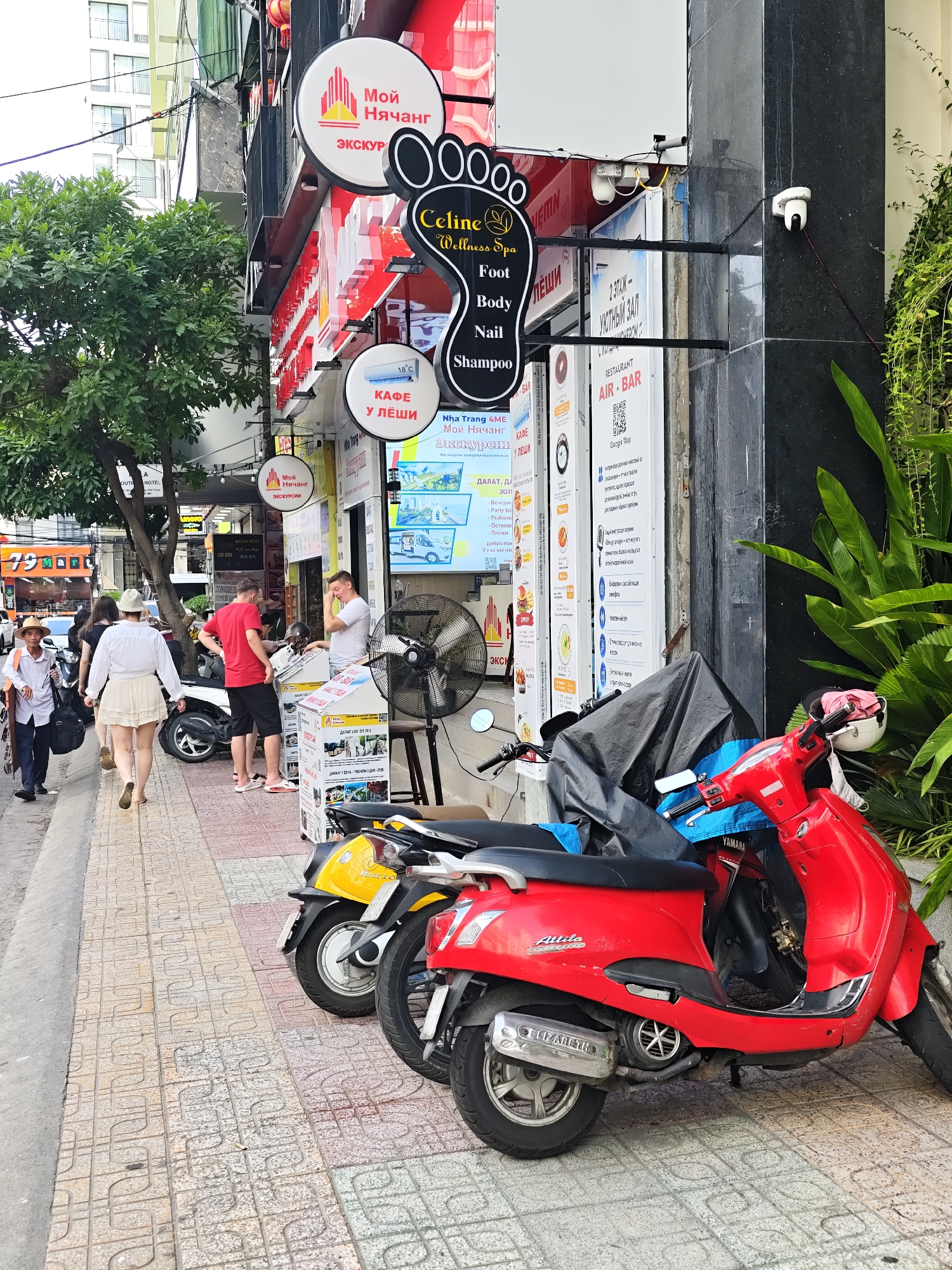 The image captures a street scene in Nha Trang Ward, Vietnam. In the foreground, two scooters are parked on a tiled sidewalk. The scooter on the right is red and branded "Attila". The scooter to its left is yellow and partially obscured. A black tarp covers another scooter.
Several people are present on the sidewalk. An elderly man in a straw hat and white shirt walks from left to right. Behind him, a woman in a white hat and dress walks away from the camera, followed by another woman in black. Further back, two men are engaged in conversation near a white scooter. One man wears a red shirt and black shorts, and the other wears a white t-shirt. A large industrial fan stands to the right of the men.
The background features a storefront with various signs. Prominent signs include a circular sign with a red and yellow design and Russian text that appears to be "Мой Нячанг" (My Nha Trang) and "Экскурсии" (Excursions). Another circular sign displays a footprint graphic and text "Celine Wellness Spa" along with "Foot Body Nail Shampoo". Several other signs with Russian text and graphics are visible, suggesting a tourist-oriented business. A security camera is mounted on the building's facade. Lush green foliage is visible to the right of the building. The overall environment appears to be a busy urban street.