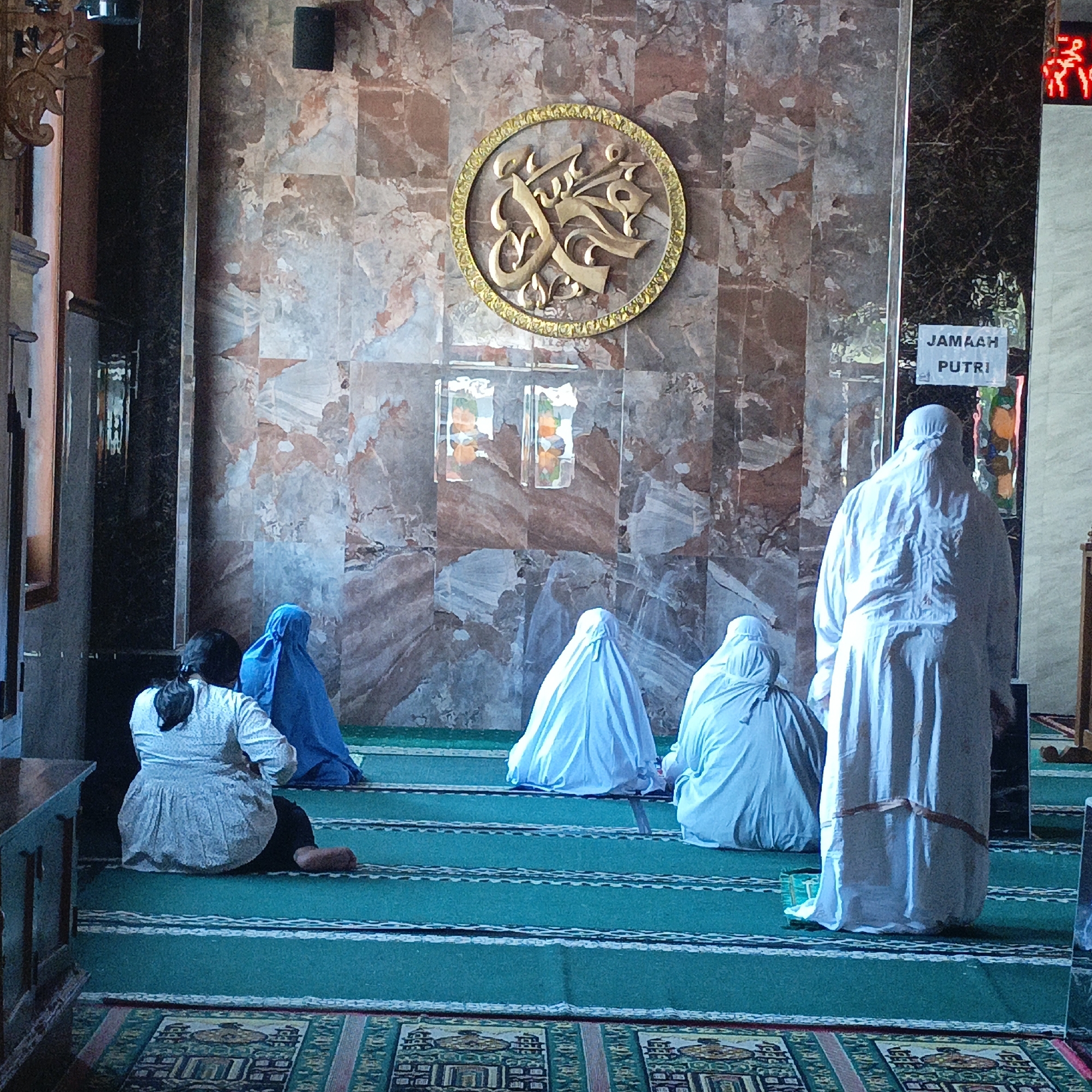 The image depicts the interior of a mosque, specifically a prayer area for women, as indicated by the sign "JAMAAH PUTRI" (Women's Congregation) visible on a pillar. The mosque features a patterned marble wall, a circular golden Islamic calligraphy emblem, and green prayer mats with white striped borders on the floor. Several women are present, all wearing traditional Islamic attire covering their heads and bodies. One woman in the foreground on the left is sitting, wearing a white patterned top and dark lower garment, facing away from the camera. To her right, another woman is seated, wearing a bright blue hijab. Further back, three other women are also seated or kneeling on the prayer mats, all covered in light blue or white attire, facing towards the wall with the calligraphy. The lighting suggests it is daytime, and the overall atmosphere appears serene and prayerful. The location is identified as Salatiga, Indonesia.
