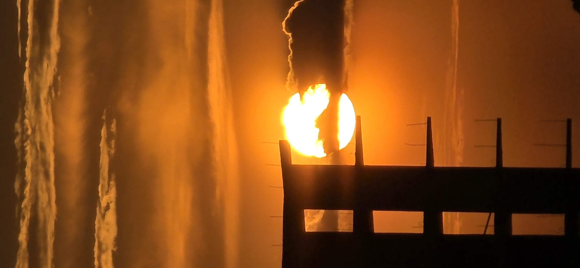 The image appears to depict a fire or explosion in Bar, Montenegro, viewed through a fence or building structure. A bright orange sun is partially obscured by smoke and flames, casting a dramatic silhouette of the structure in the foreground. Light and shadow play across the scene, creating a sense of intensity and danger. Long vertical streaking marks cover left half of the image suggesting heavy rain.