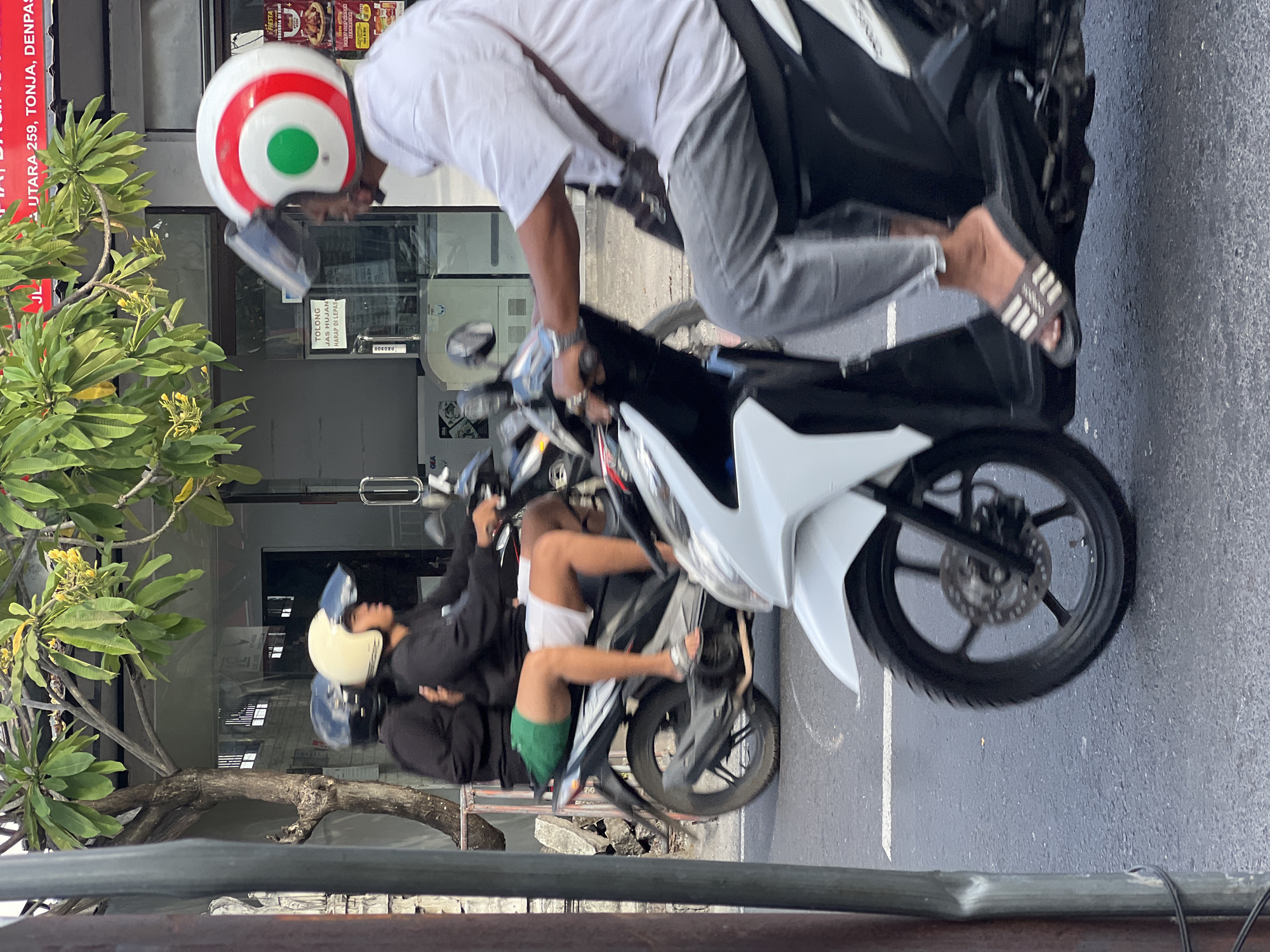 A man wearing a white short-sleeved shirt, grey pants, and flip-flops rides a white scooter. He has a brown bag slung across his chest. A second scooter is riding in front of him, with two people on it. The person in front is wearing a white helmet and a black shirt, and the person in the back is wearing a black helmet and a black shirt with green shorts. The scooters are on a paved road in Denpasar, Indonesia. A tree with green leaves and yellow flowers is visible in the background. Buildings with signage are also visible in the background.