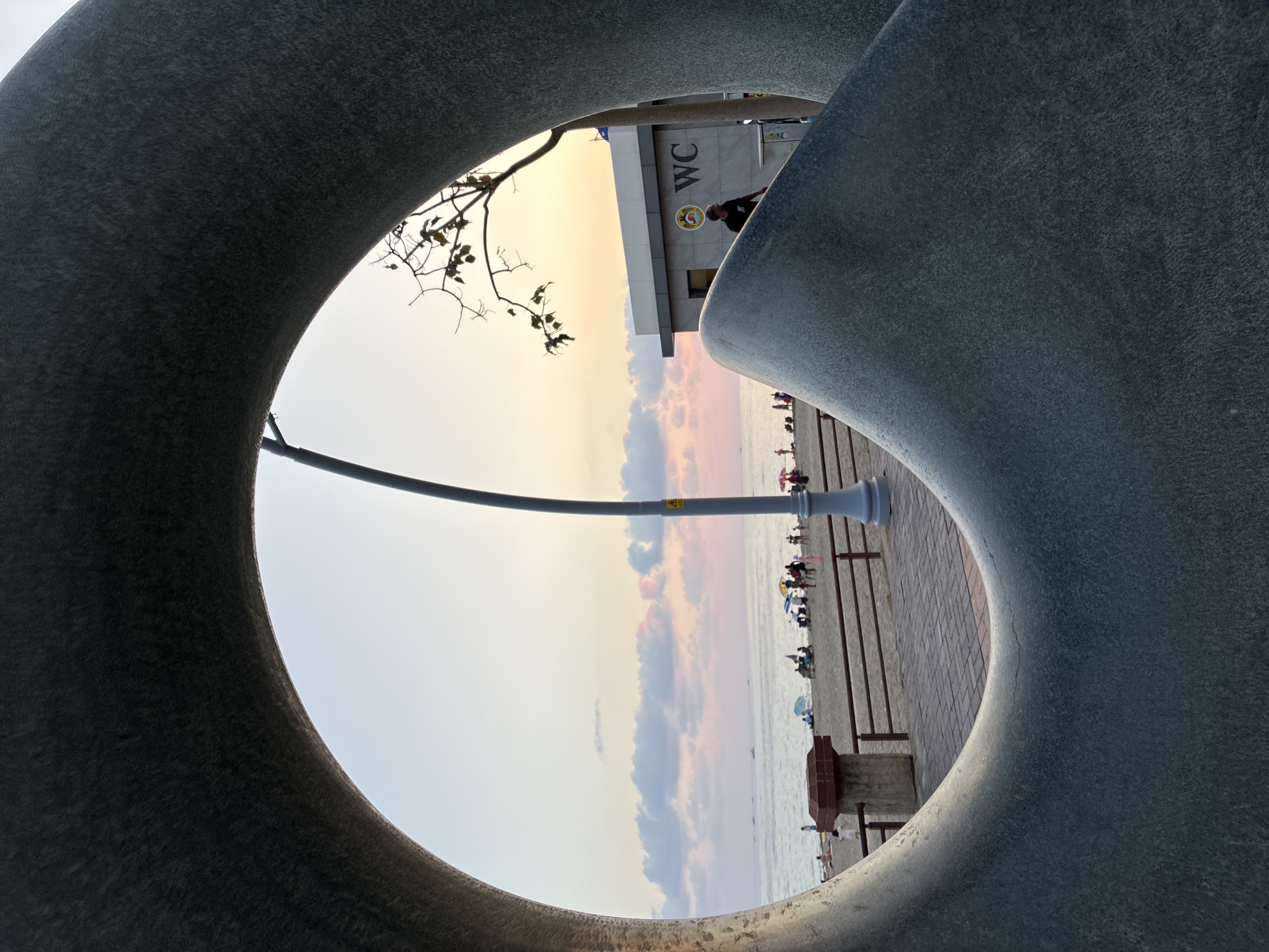 The image shows a view through a large, circular, abstract sculpture. The scene beyond the sculpture is a beach with people relaxing on the sand and in the water. The sky is a soft gradient of pink, orange, and blue, indicating either sunrise or sunset. In the background, there is a building with a "WC" sign, suggesting it's a public restroom facility, likely located near the beach. A lamppost stands near the building. The sculpture itself is made of a rough, textured material, possibly stone or concrete, and appears to be a prominent art installation on the Alanya promenade, a popular tourist area in Alanya, Turkey.
