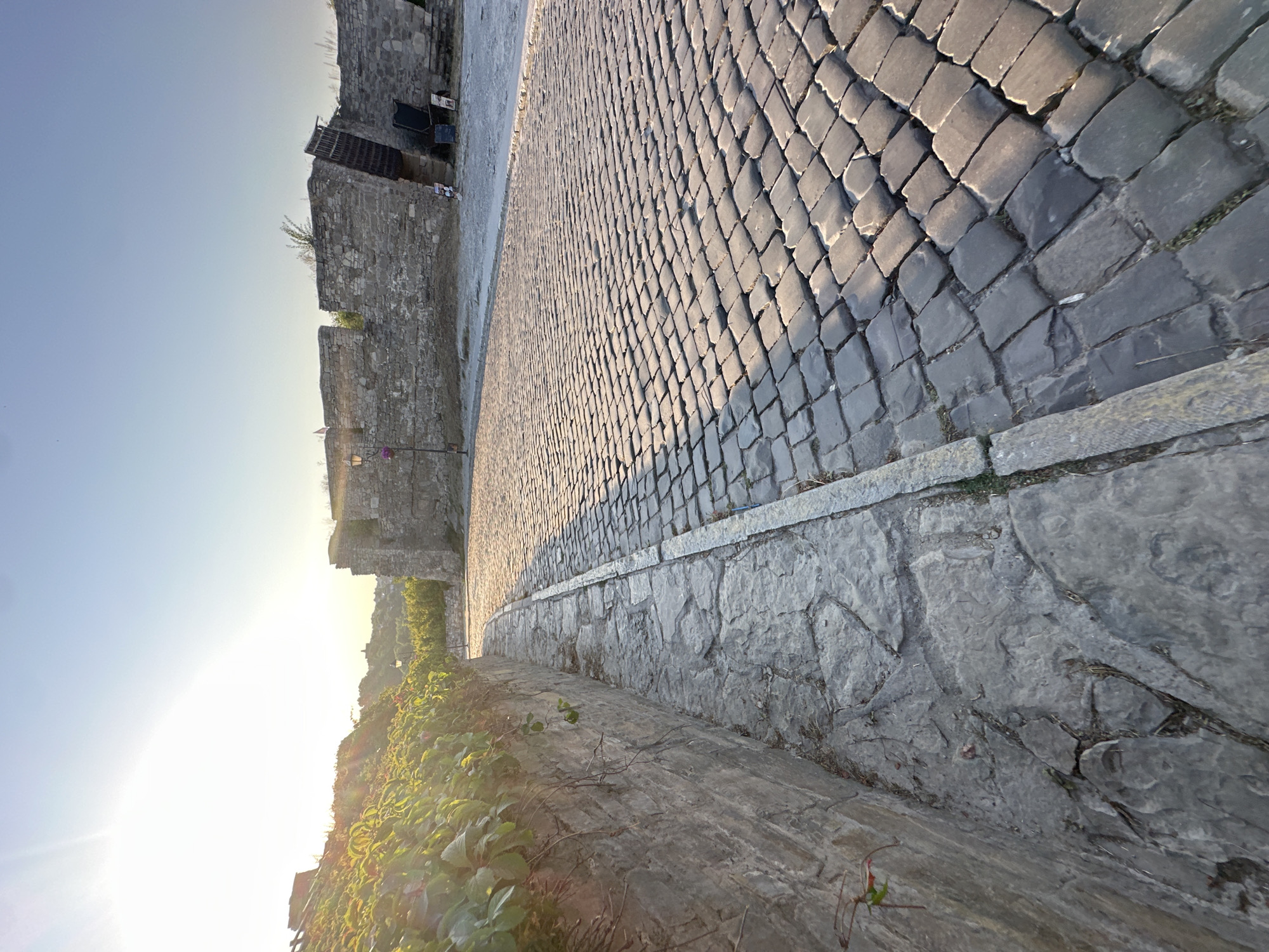 The image shows a cobblestone street leading towards the walls of a fortress in Kamianets-Podilskyi, Ukraine. The sun is setting, casting a warm glow on the scene. The street is paved with dark, irregularly shaped cobblestones. To the left of the street, a stone wall is covered with lush green foliage. To the right, the fortress walls, made of rough-hewn stones, stand tall with battlements. The sky is clear and blue, with a bright sun creating a lens flare in the upper left corner. There are no people visible in the image. The scene evokes a sense of history and timelessness.