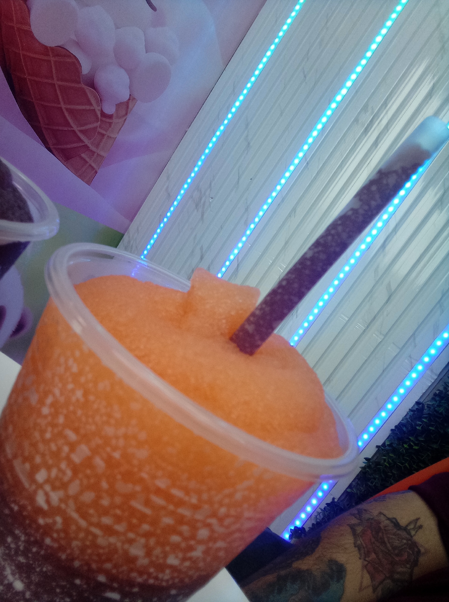 A close-up, high-angle shot shows a clear plastic cup filled with an orange slushie-type drink. A brown-spotted straw is inserted in the drink. In the background, there is a white wall with vertical blue LED strips and a poster of an ice cream cone. Partial view of a person's arm with a tattoo is visible in the lower right corner. The location may be a commercial establishment in Guarenas, Venezuela.