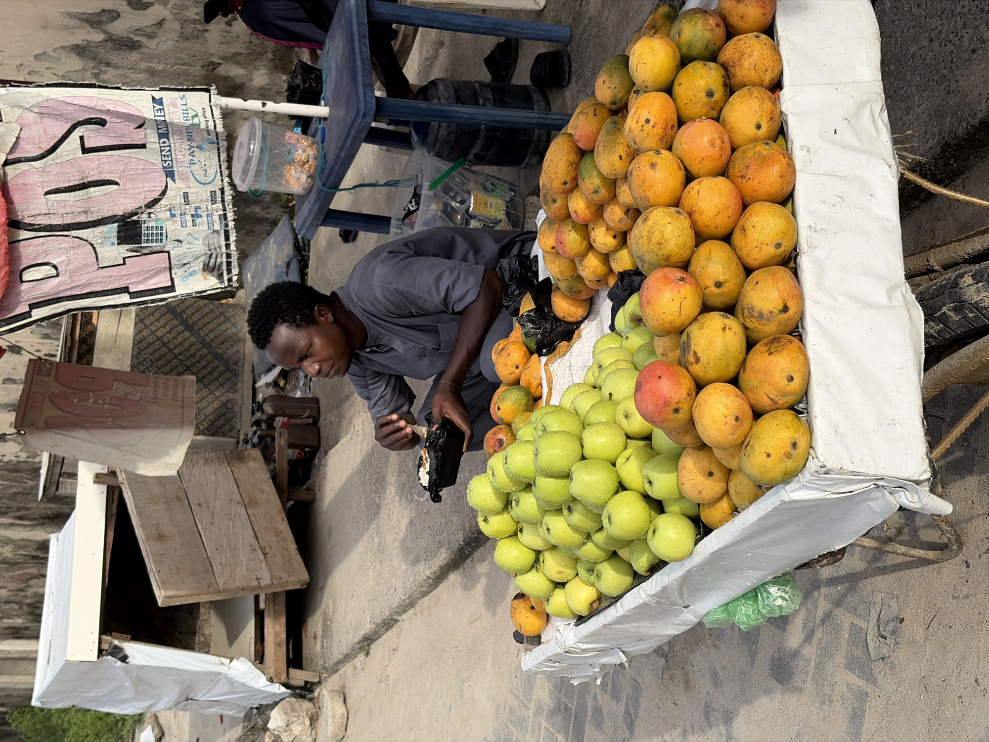 The image captures a vibrant street market scene in Igboefon, Nigeria, under bright daylight. The main subject is a man, likely a vendor, who is engaged in tending to his fruit stall. He is dressed in a grey shirt and is positioned on the ground, seemingly focused on handling items within a black plastic bag.
The foreground is dominated by a prolific display of fresh produce arranged on what appears to be a makeshift stall covered with white fabric. A large pile of yellow and reddish-orange mangoes is prominently stacked, alongside an equally large mound of bright green apples or guavas, some of which appear to have water droplets on them, suggesting freshness.
In the background, various elements of a bustling local environment are visible. To the left of the vendor, there is a blue plastic table and chairs, and a clear plastic container holding snacks or other small goods. Faded wooden structures and shelving indicate a simple, possibly temporary, market setup.
Several pieces of text are discernible throughout the scene. A large, weathered banner or sign hangs above, bearing the prominent phrases "SEND MONEY" and "PAYNE BILLS," with "hing africa" also visible, suggesting a financial transaction service. Elsewhere, text such as "PROSSY SUPER QUALITY," "SUPER QUALITY," and "SUPER QU" is repeated on what appears to be packaging or another label. The numbers "30000" can also be seen. The overall atmosphere conveys a sense of daily commerce and local activity.
