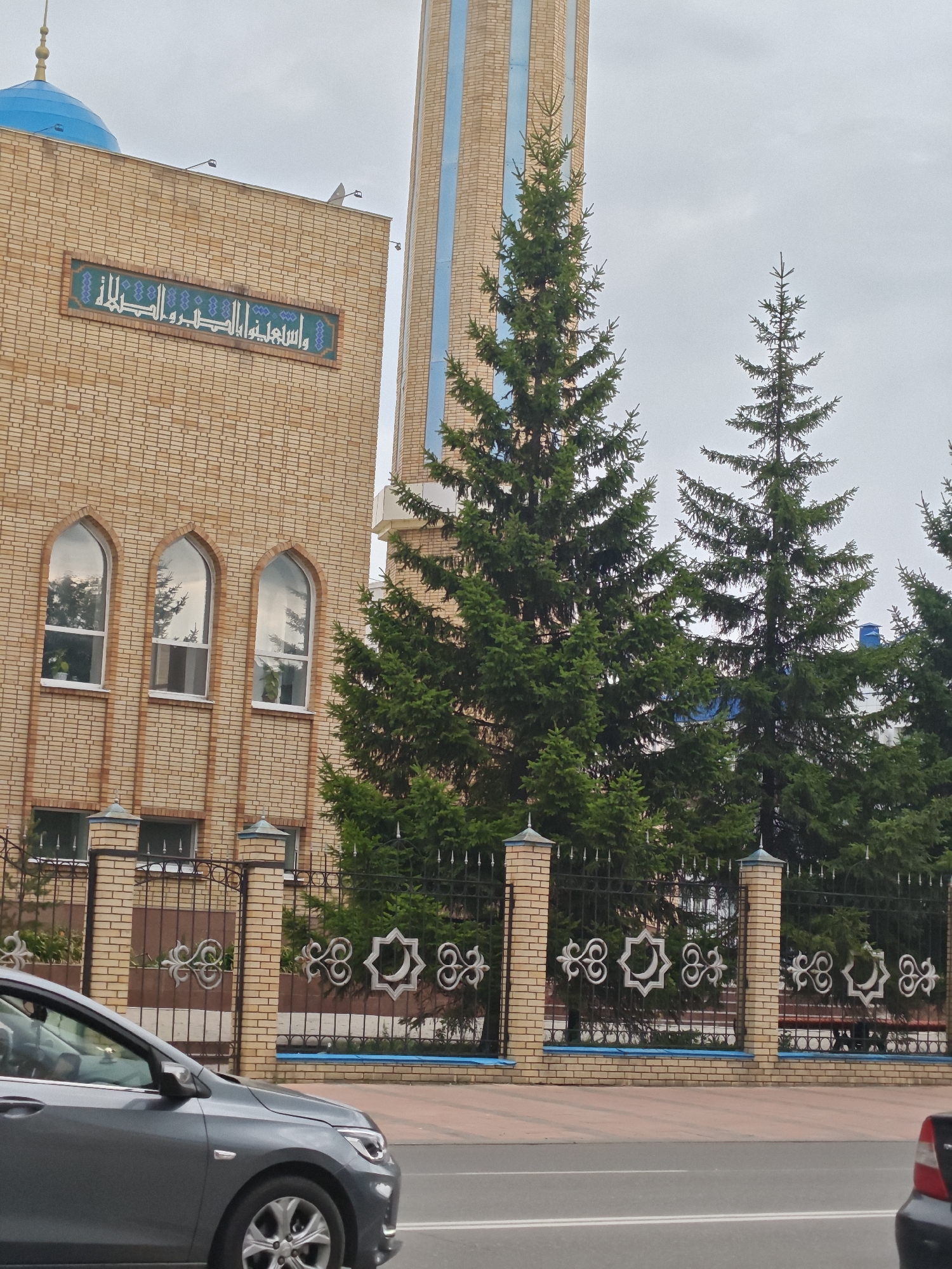 A two-story mosque with a blue dome and minaret is visible in Petropavl, Kazakhstan. The mosque is made of light brown brick and features arched windows. The exterior is decorated with an Arabic inscription and intricate metalwork on the fence. Several evergreen trees are planted in front of the mosque, and a grey car is driving on the street in the foreground. The sky is overcast.