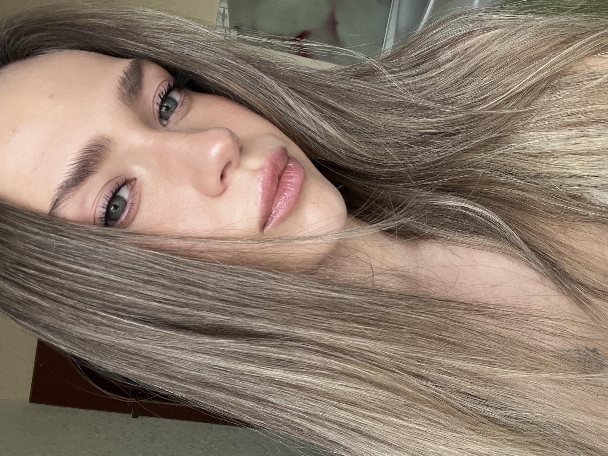 A head-and-shoulders portrait of an adult female is depicted indoors. The specific location within Khimki, Russia, is not discernible from the visual information. The subject has long, straight, ash-blonde hair that falls around her face and over her shoulders. Her head is slightly tilted, and her gaze is directed towards the camera with a neutral facial expression. Her eyes are light-colored, and she has full, well-defined eyebrows and prominent eyelashes. Her lips are full and appear glossy with a light pink tint. Her skin appears smooth. The background is out of focus, revealing indistinct elements: a dark brown vertical object, possibly a door frame, against a light-colored, textured wall to the left, and a lighter, indistinct, possibly reflective surface to the right. The individual is stationary, posing for the photograph.