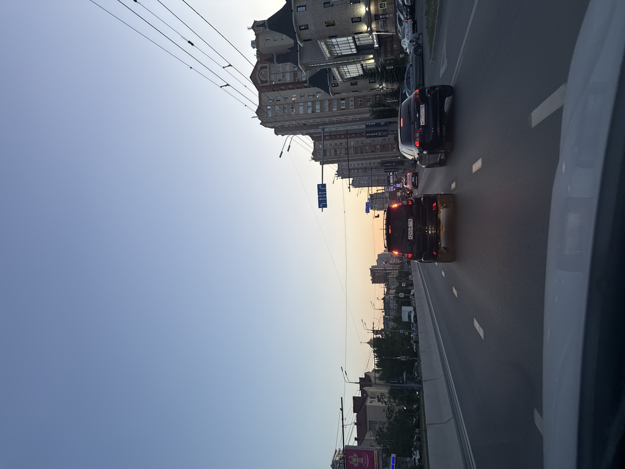The image depicts a street scene in Kazan, Russia. The view is from inside a car, looking along a multi-lane road. Several vehicles are visible in the lanes ahead, including two cars with license plates K245HK716 and M722AT116. Buildings line the street, including a tall building with distinctive architectural features. Streetlights and traffic signals are present. The sky appears to be late in the day, possibly dusk, indicated by the light.