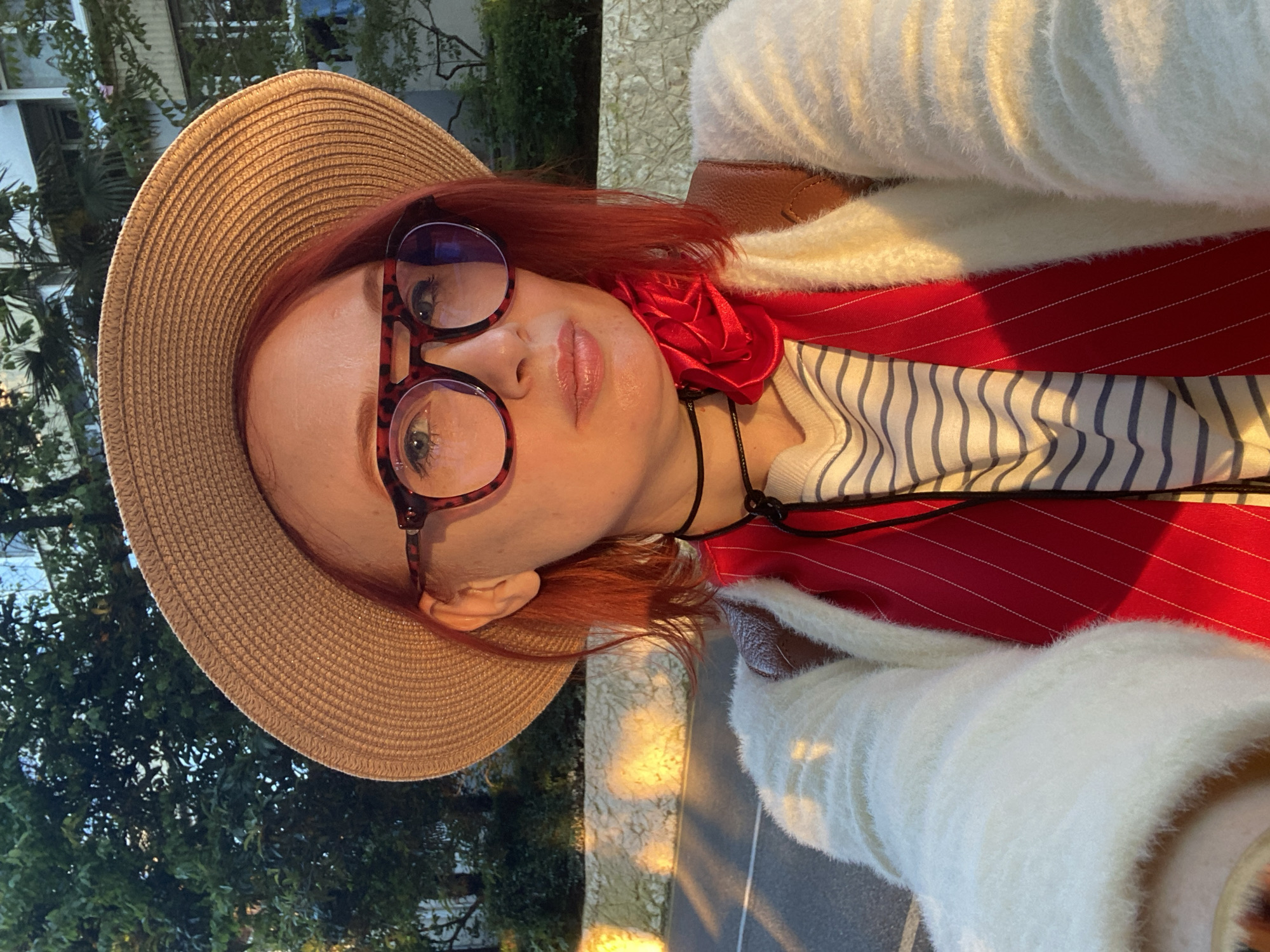A close-up, rotated selfie features an individual with reddish-orange hair, wearing a light brown woven straw hat and large, round tortoiseshell eyeglasses. A red fabric flower is pinned near their left ear. They are dressed in a white fluffy sweater, layered over a red pinstriped jacket or blazer, and an inner striped shirt. A black choker necklace is visible. The person has a neutral to slightly pensive expression, looking directly at the camera.
The background shows lush green trees and foliage, suggesting an outdoor setting. Warm, golden sunlight illuminates parts of the scene, casting bright highlights on the hat, face, and the ground, indicating either early morning or late afternoon. The overall impression is a bright, sunny day. The location context is Sochi, Russia.