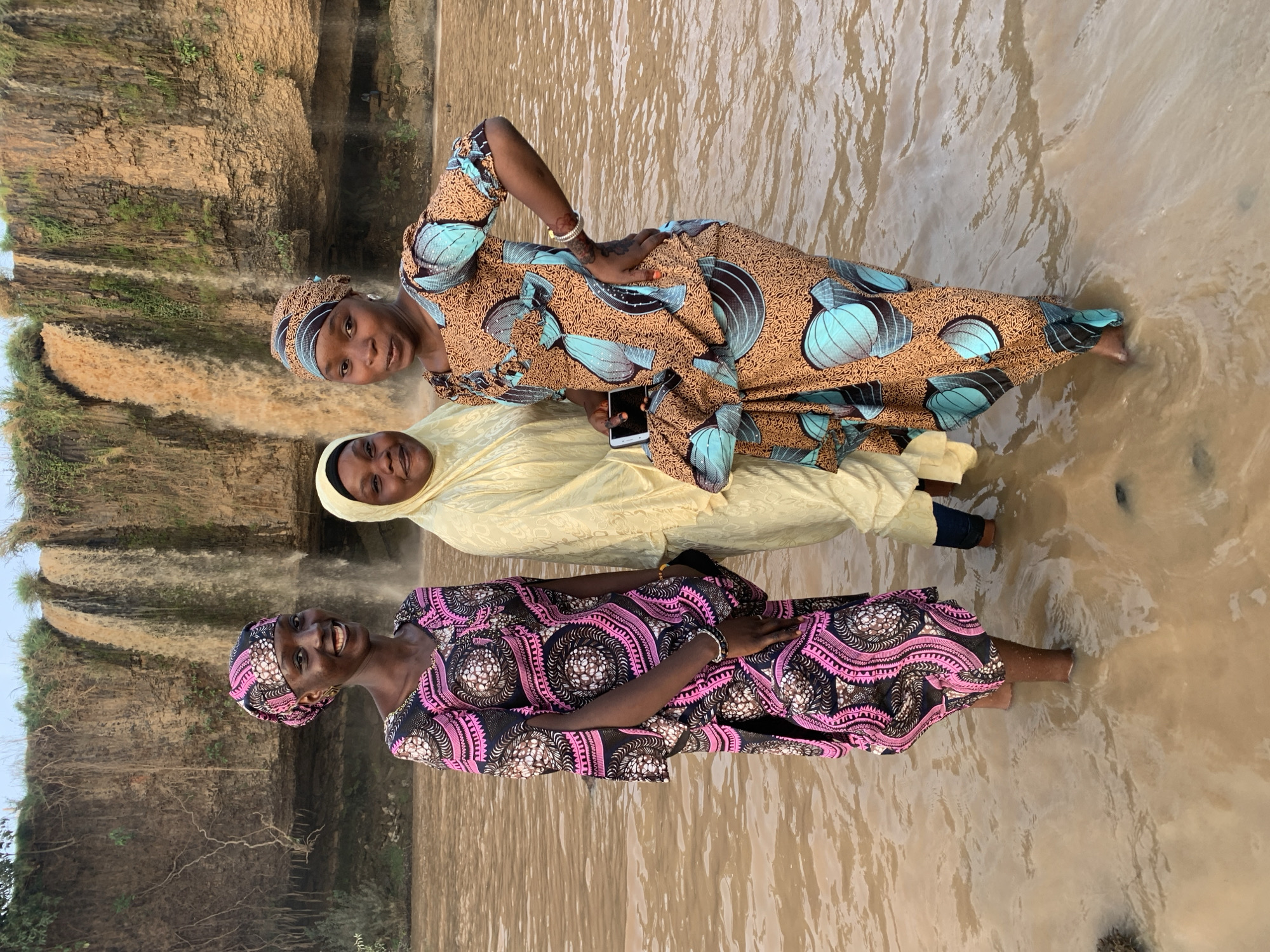 Three women stand together in shallow water at the base of a waterfall. The woman in the foreground wears a pink and black patterned dress with a matching head wrap and smiles at the camera. The woman in the middle wears a light yellow dress and head covering. The woman in the back wears a brown dress with blue and black patterns and also smiles at the camera, holding a cell phone. The water is muddy. The waterfall is visible behind them, cascading down a rocky cliff. The scene is in Kukum, Nigeria.