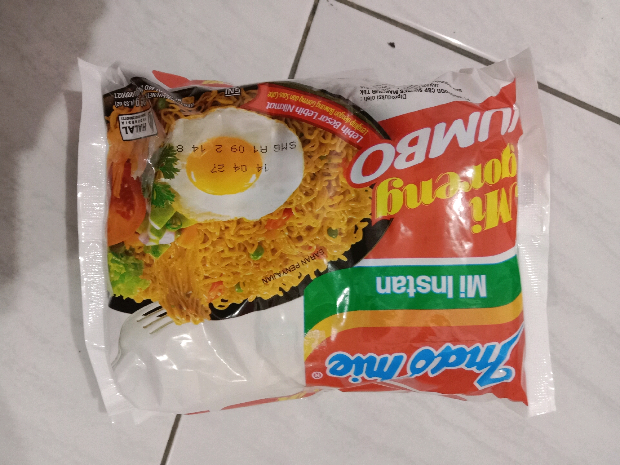 A top-down, medium shot shows a package of Indomie Mi Goreng Jumbo instant noodles on a tiled floor. The packaging is predominantly red and white, with the brand name "Indomie" in blue script on the bottom half. The top half features a close-up image of a fried egg atop a serving of noodles, with some vegetables visible. Text in Indonesian and English is present on the packaging, including "Mi Instan Jumbo" and "Lebih Besar Lebih Nikmat" (Bigger is More Delicious). A small "HALAL" logo is visible on the left side of the packaging. The tiled floor appears to be white with grey grout lines. There are no people or other objects visible in the frame. The image is lit from above, casting soft shadows.