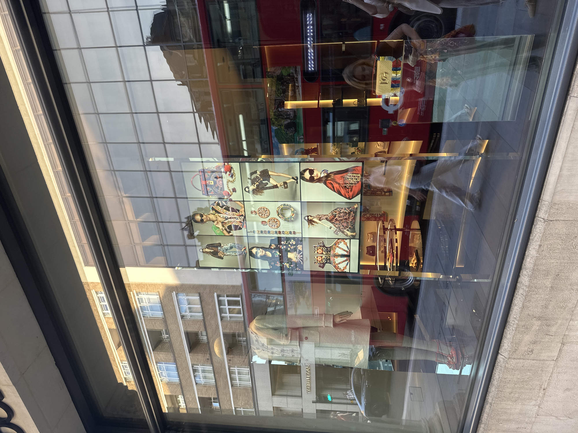 The image shows a street-level view of a store window. Reflected in the window is a red double-decker bus, a brick building with white-framed windows, and part of another building with a more modern facade. Inside the store, a mannequin is displayed wearing a light-colored tweed suit and red high-heeled shoes. To the right of the mannequin, a series of digital screens show fashion imagery of models wearing stylish outfits and accessories, along with close-ups of bags and jewelry. In the background of the store, shelves are stocked with handbags.
Outside the store, a black car is parked on the street, and several pedestrians are visible walking on the sidewalk. One woman, partially obscured, is looking at her phone. Another person is walking in the foreground, their silhouette cast on the floor. The lighting suggests it is daytime, with sunlight casting shadows and highlighting reflections on the glass. The overall scene depicts a fashion retail environment in an urban setting.