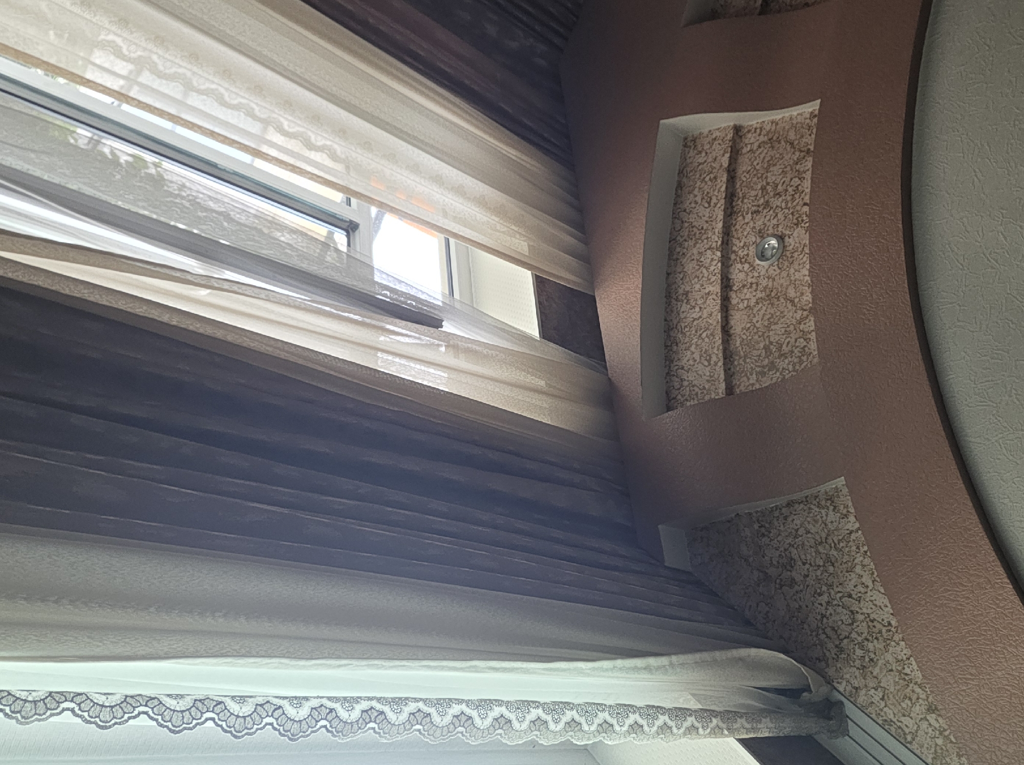 A low-angle shot shows a multi-layered window treatment in a room. The ceiling features a decorative plaster design with textured panels and a recessed light fixture. The window is covered by several sheer curtains in varying shades of beige and brown, and a white lace-trimmed curtain on the right. The curtains are drawn, partially obscuring a view of the window and the sky outside. The room appears to be a residential interior. The location is the city of Kirov, Kyrgyzstan.