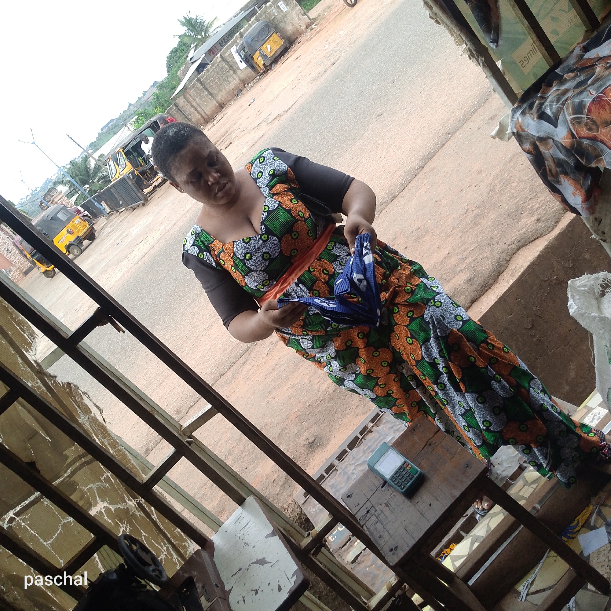 A woman stands in a shop in Nsugpe, Nigeria. She is wearing a colorful, patterned dress with black sleeves and an orange sash, and holding a folded blue object. The shop's interior is cluttered with various items and a weathered table holding a calculator. Outside, there is a street with motor tricycles ("keke napeps") visible in the distance. The perspective of the shot is angled, with a corner of the shop frame visible in the foreground.