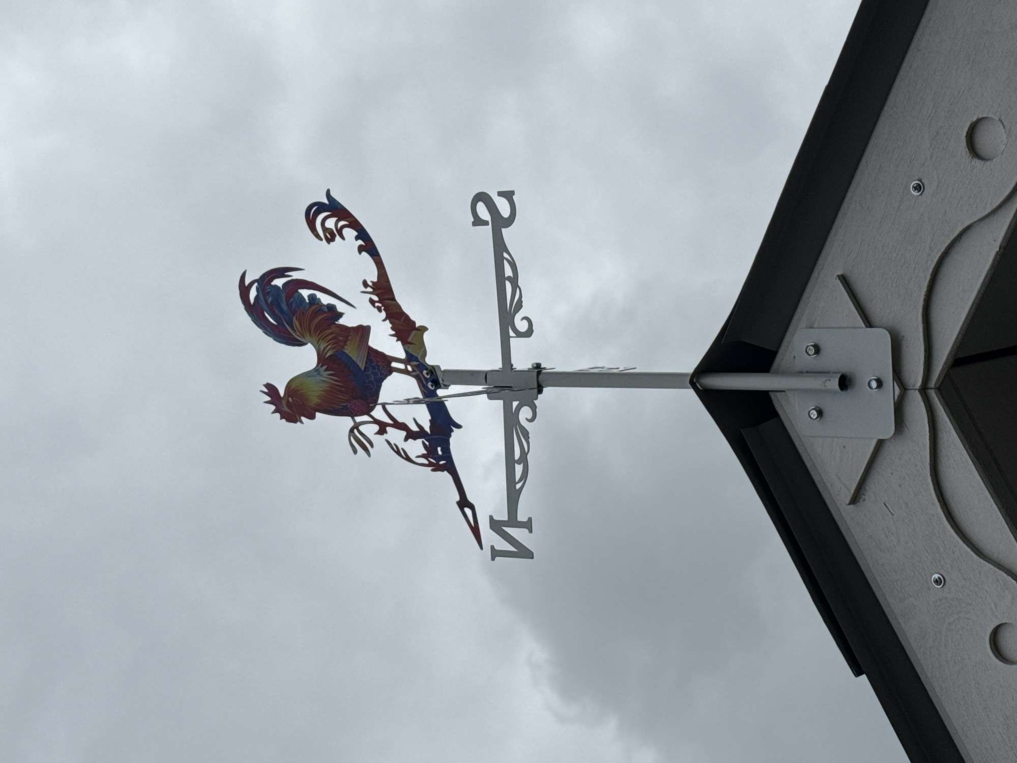 The image depicts a colorful rooster weathervane mounted on the roof of a structure. The weathervane features a rooster figure and directional indicators oriented towards north, south, east, and west. The background consists of a cloudy sky and the edge of the building structure. The location is presumed to be Chaikovsky, Russia.