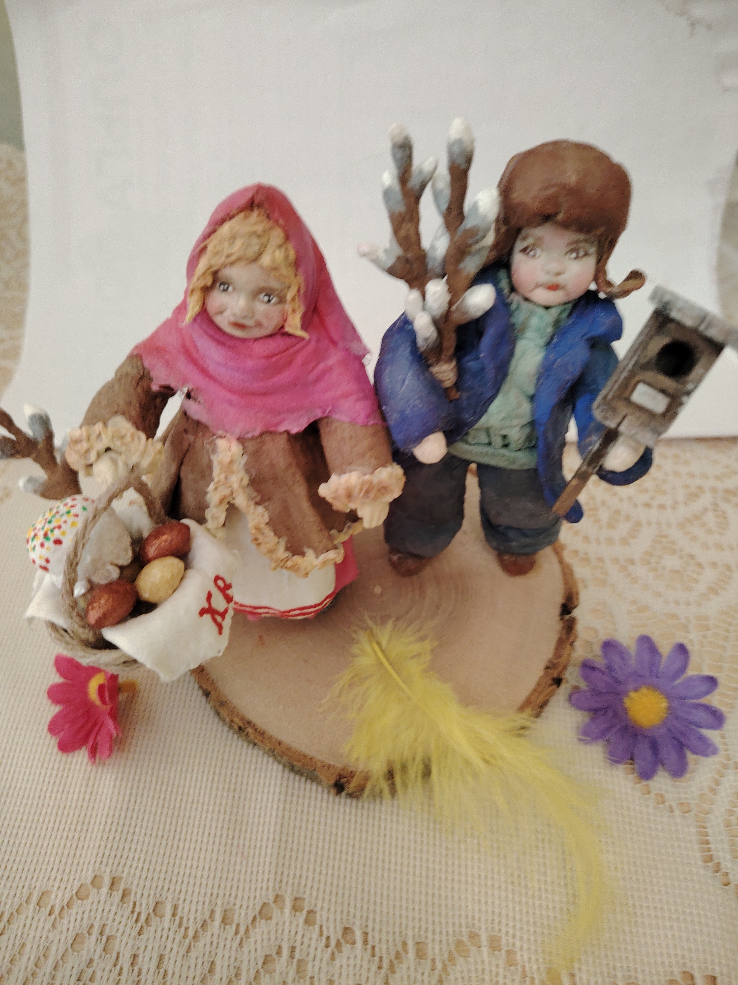 A close-up studio shot depicts two handcrafted, painted figurines on a wooden base, surrounded by artificial flowers and a feather. The figure on the left wears a pink headscarf and a brown dress with a white apron bearing red Cyrillic characters, carrying a basket of eggs and sprigs of pussy willow. The figure on the right wears a blue coat and dark pants, holding pussy willow branches and a small birdhouse. The artificial flowers are pink and purple, and a yellow feather lies nearby. The background is blurred white. The figurines and the arrangement evoke themes of Easter or spring, potentially of Russian or Eastern European origin. This location is not stated in the image.