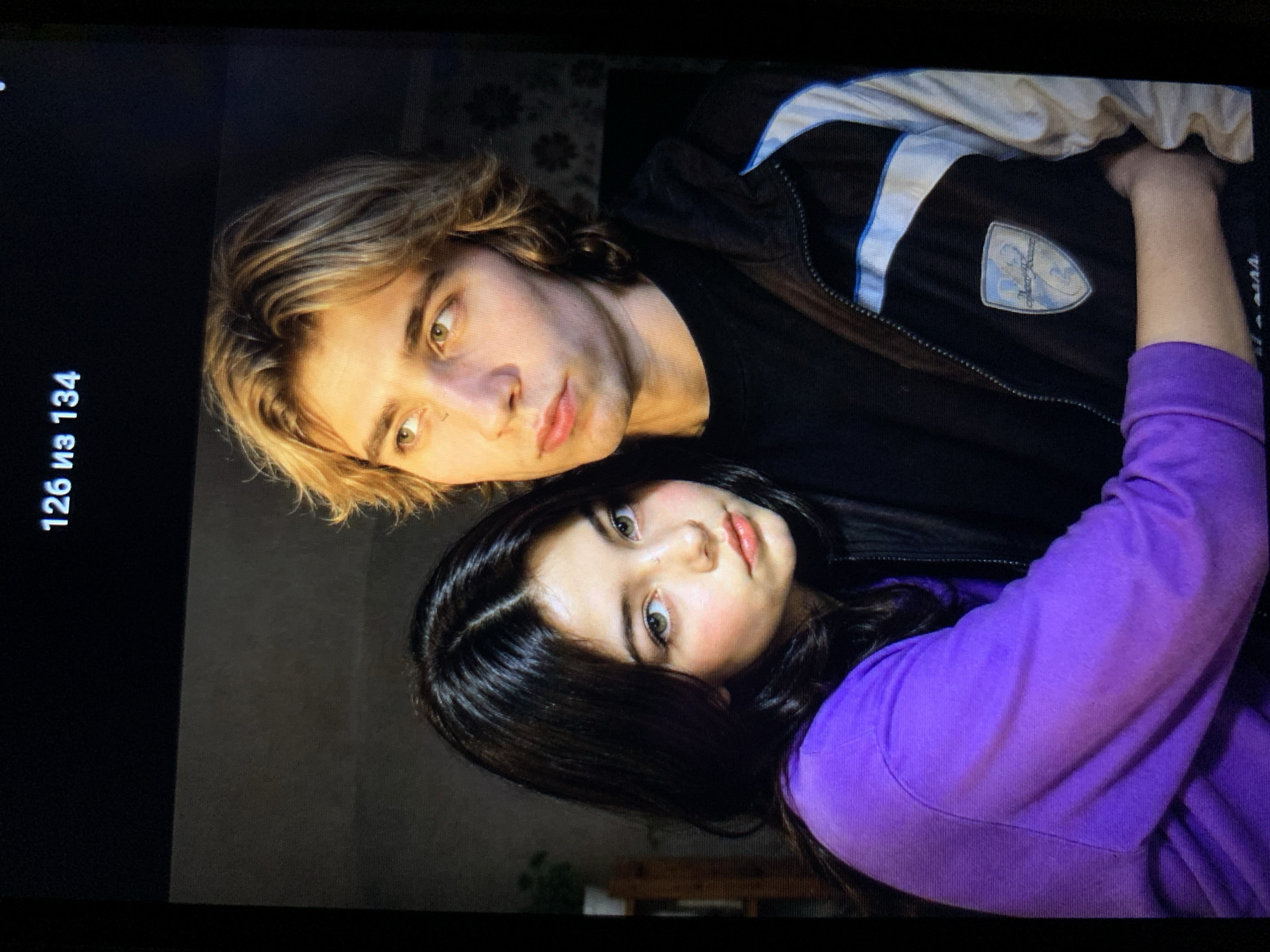 The image shows a close-up of a young couple. The woman on the left has long, dark hair and is wearing a purple hoodie. Her skin appears to have a light blush on her cheeks, and her eyes are looking towards the viewer. The man on the right has medium-length blond hair and is wearing a black sweatshirt under a dark jacket with white and blue accents. He is looking away from the camera. They are positioned close together, with the woman leaning into the man. The background is dark and indistinct, with some hints of a room interior, including a shelf with a plant on the left and patterned wallpaper on the right. The text "126 из 134" is visible in the upper right corner of the image, indicating page numbers. The provided image does not contain any discernible elements that would allow for its identification to a specific location within the city of Lymanka, Ukraine.