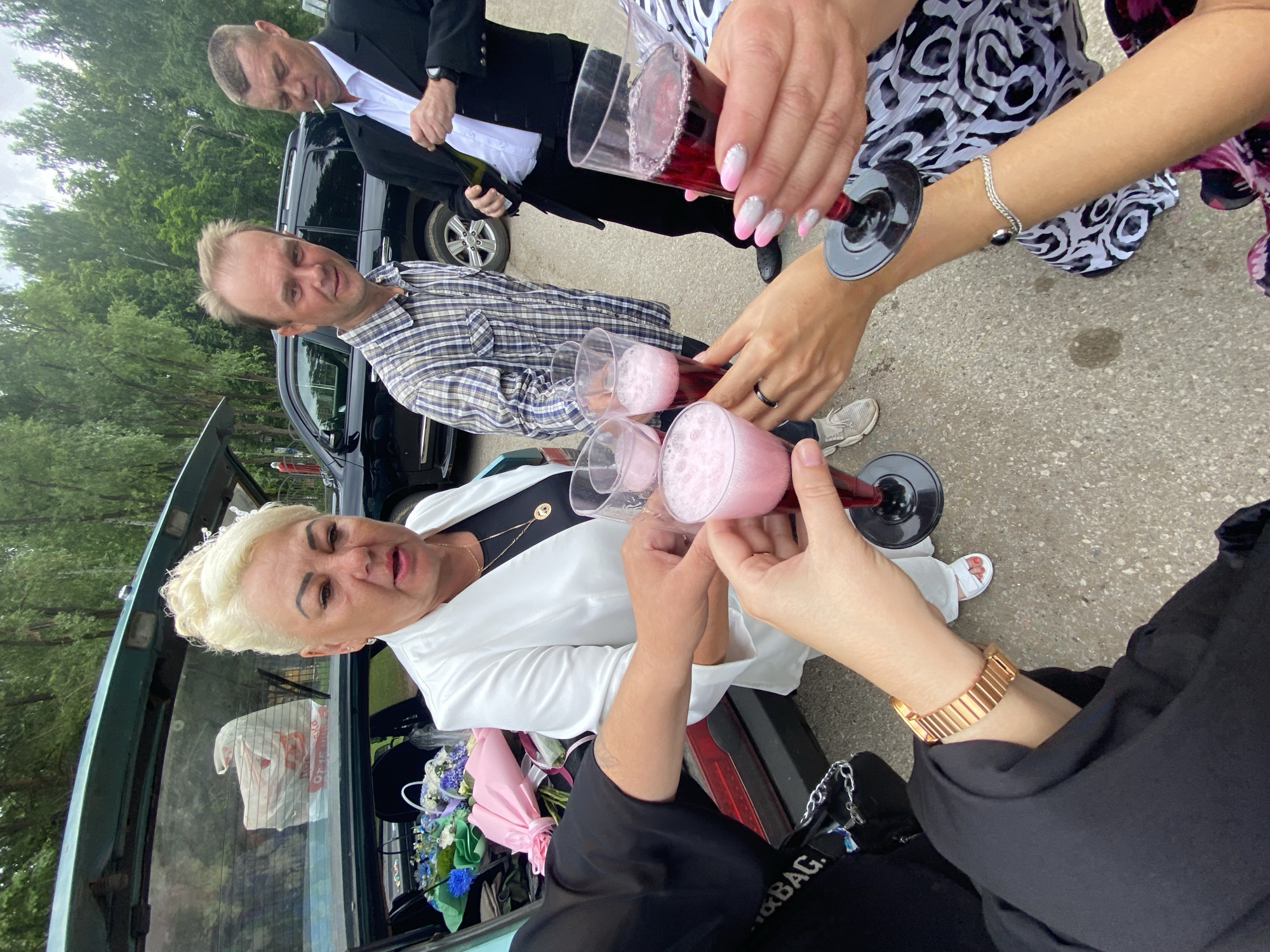 In Chaikovsky, Russia, a group of four adults is gathered outdoors, near an open car trunk. One woman, with blonde, up-swept hair and wearing a white blazer over a black top, is in the foreground, holding a pink beverage in a clear plastic flute. To her right, a man in a plaid shirt is looking at the camera, while another man in a black suit and white shirt is holding a green bottle and a cigarette. The car trunk contains several bouquets of flowers, including blue and purple arrangements, and a pink wrapped bouquet. The ground is paved with asphalt.