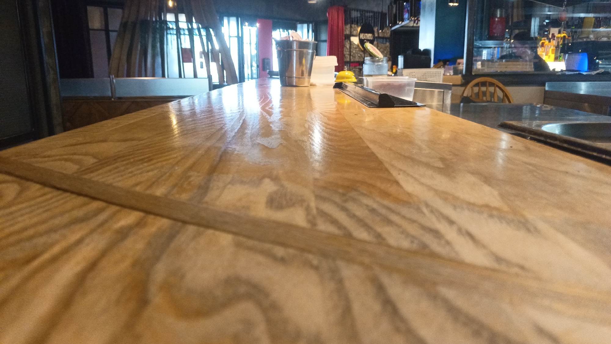 The image shows a close-up, low-angle view of a polished wooden bar counter. The counter has a light wood grain with variations in tone, showing a glossy finish with reflections. On the counter, there are several objects including a silver metal bucket filled with items, a small yellow bell, a clear plastic container, and what appears to be a black metal stand or tray. In the background, the scene suggests an indoor establishment, possibly a bar or restaurant, with elements of decor like curtains, lights, and shelves with bottles visible. The specific location in Mytishchi, Russia, cannot be determined from the visual information provided. There are no people clearly visible, and no specific actions or interactions are depicted.